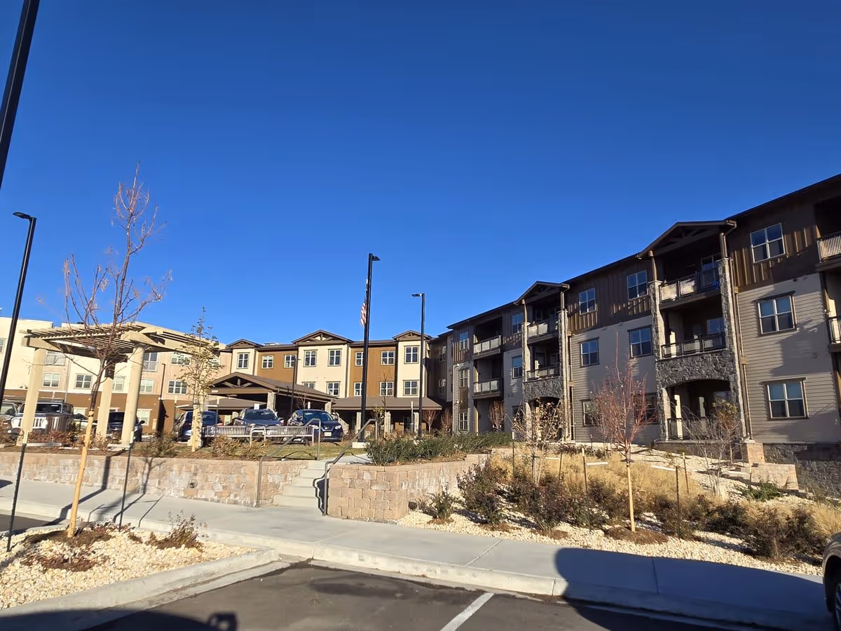 Front exterior of a multi-story senior living building with balconies, parking and landscaped grounds under a clear blue sky