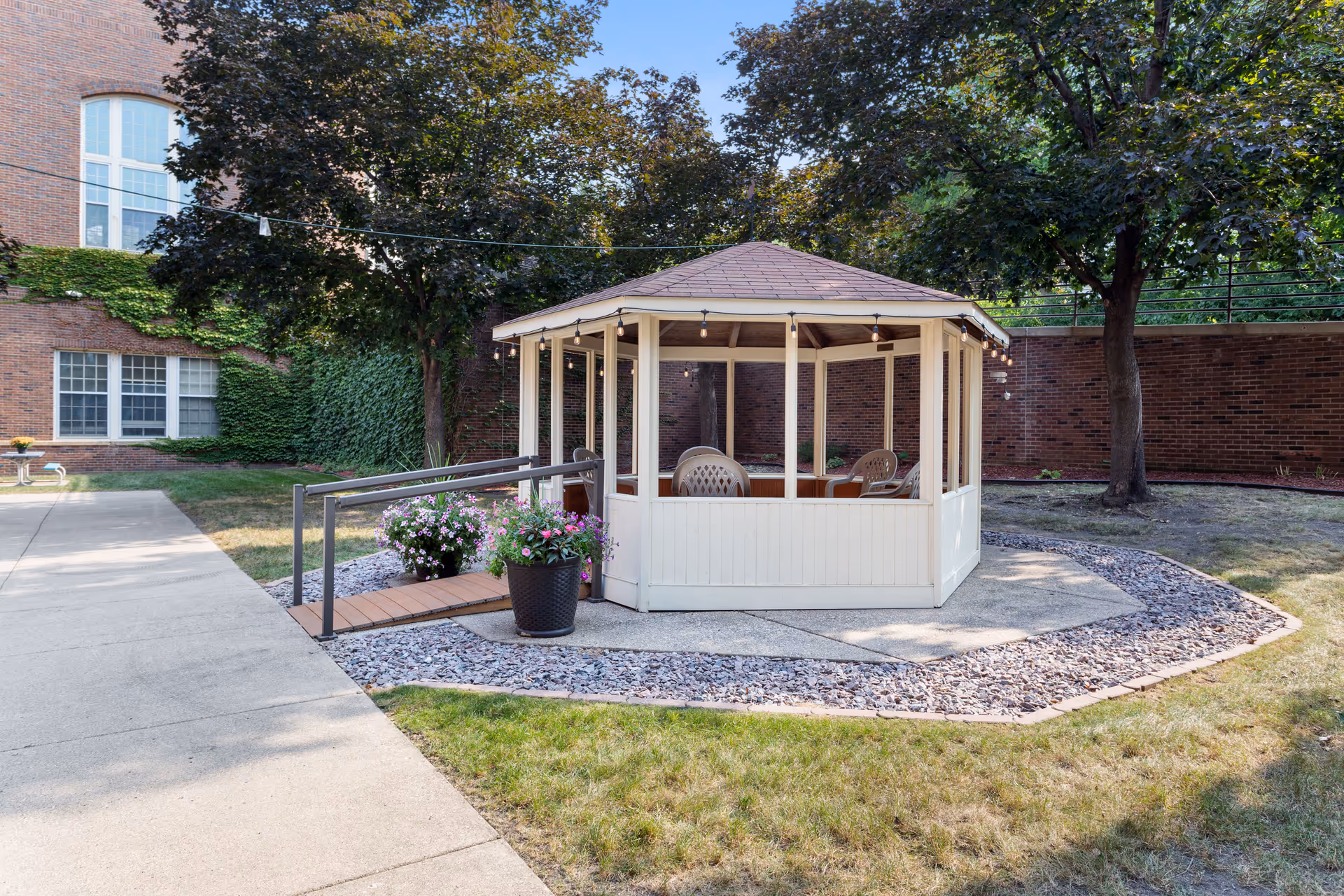A white gazebo with a brown roof situated in a garden area with trees and grass. The gazebo has plastic chairs inside and is surrounded by a stone border. There is a small wooden ramp leading to the gazebo, and potted flowers are placed near the entrance. In the background, there is a brick building partially covered with ivy.