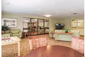 Community lounge with round tables and upholstered chairs, sofas, a TV, and glass doors opening to a dining area.