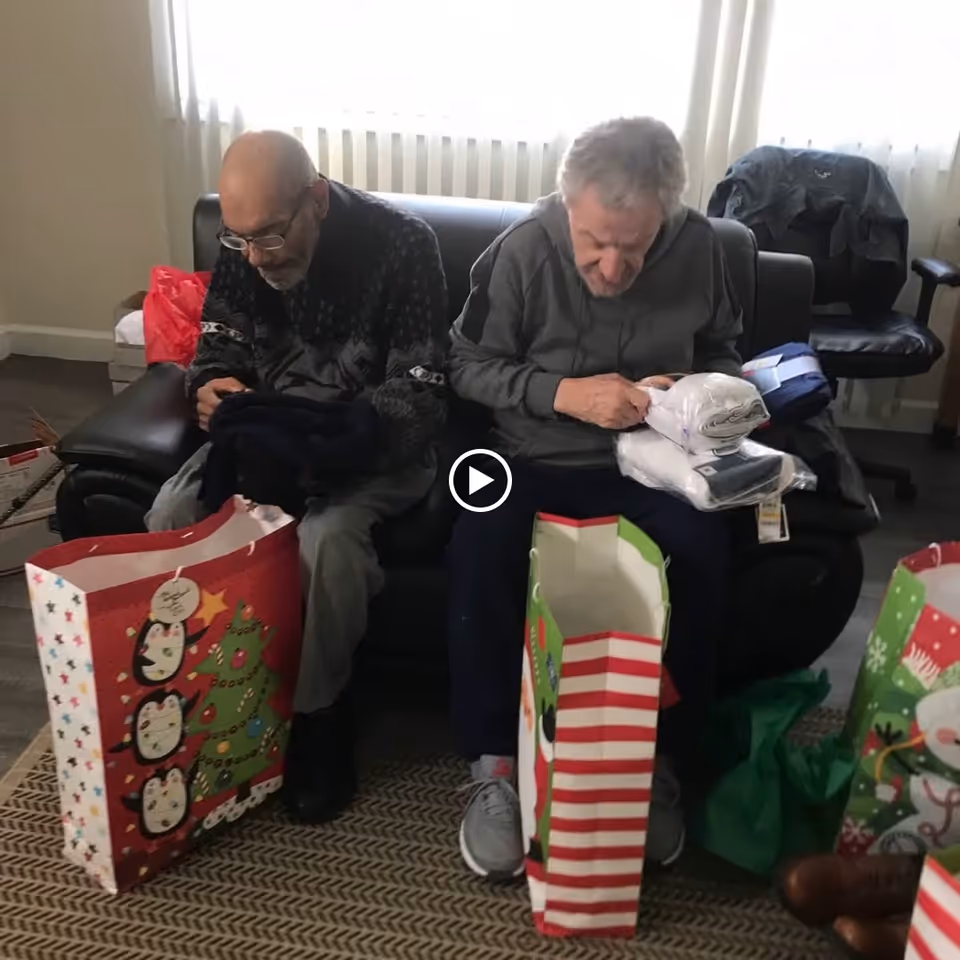 Two elderly men sitting on a black couch in a living room, each holding items they have taken out of large festive gift bags placed in front of them. The room has a window with white curtains and a patterned carpet on the floor.