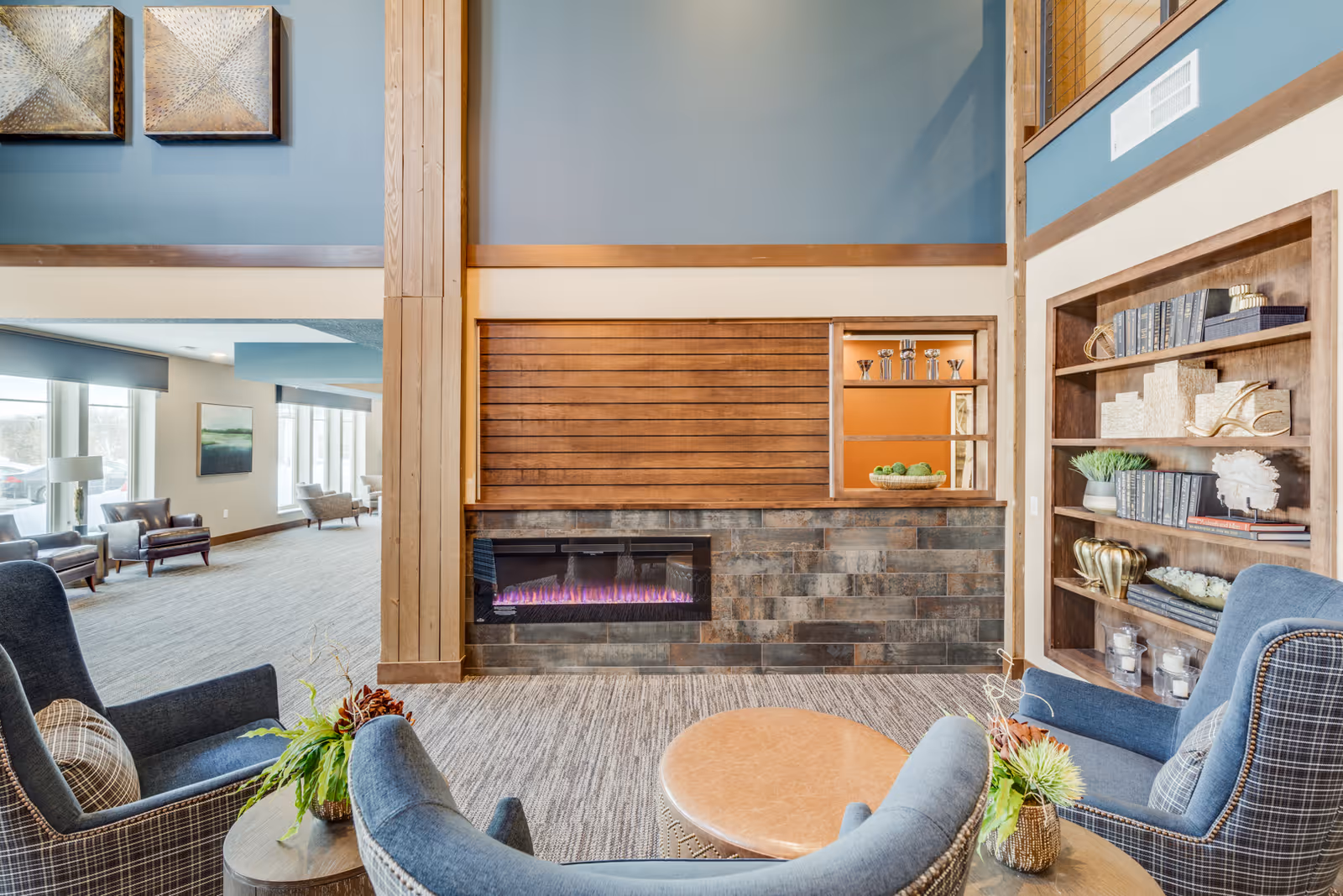 A cozy living room area in a senior living facility featuring two blue upholstered armchairs with plaid cushions, a round wooden coffee table, a modern electric fireplace with a stone surround, and built-in wooden shelves decorated with books, plants, and decorative items. Large windows in the background let in natural light, and additional seating is visible in an adjacent room.