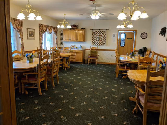A cozy dining room with multiple wooden tables and chairs arranged neatly. The room has green carpet with a floral pattern, cream-colored walls, and several ceiling lights with lampshades. There are windows with floral curtains on the left wall, wooden cabinets on the far wall, and a door with a window on the right side. Decorative items such as a quilt hanging on the wall and small plants are also visible.