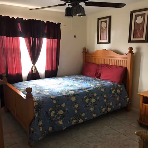 A bedroom with a wooden bed frame and headboard, a floral blue bedspread, and two red pillows. There are two framed floral pictures on the wall above the bed. A window with dark red curtains is on the left side, and a ceiling fan with lights is mounted on the ceiling. A wooden nightstand is visible on the right side of the bed.