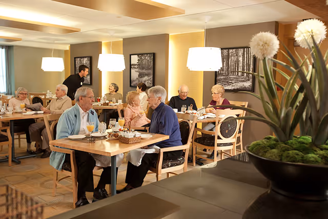 Elderly residents eating and chatting at tables in a well-lit communal dining room with pendant lamps and wall artwork.