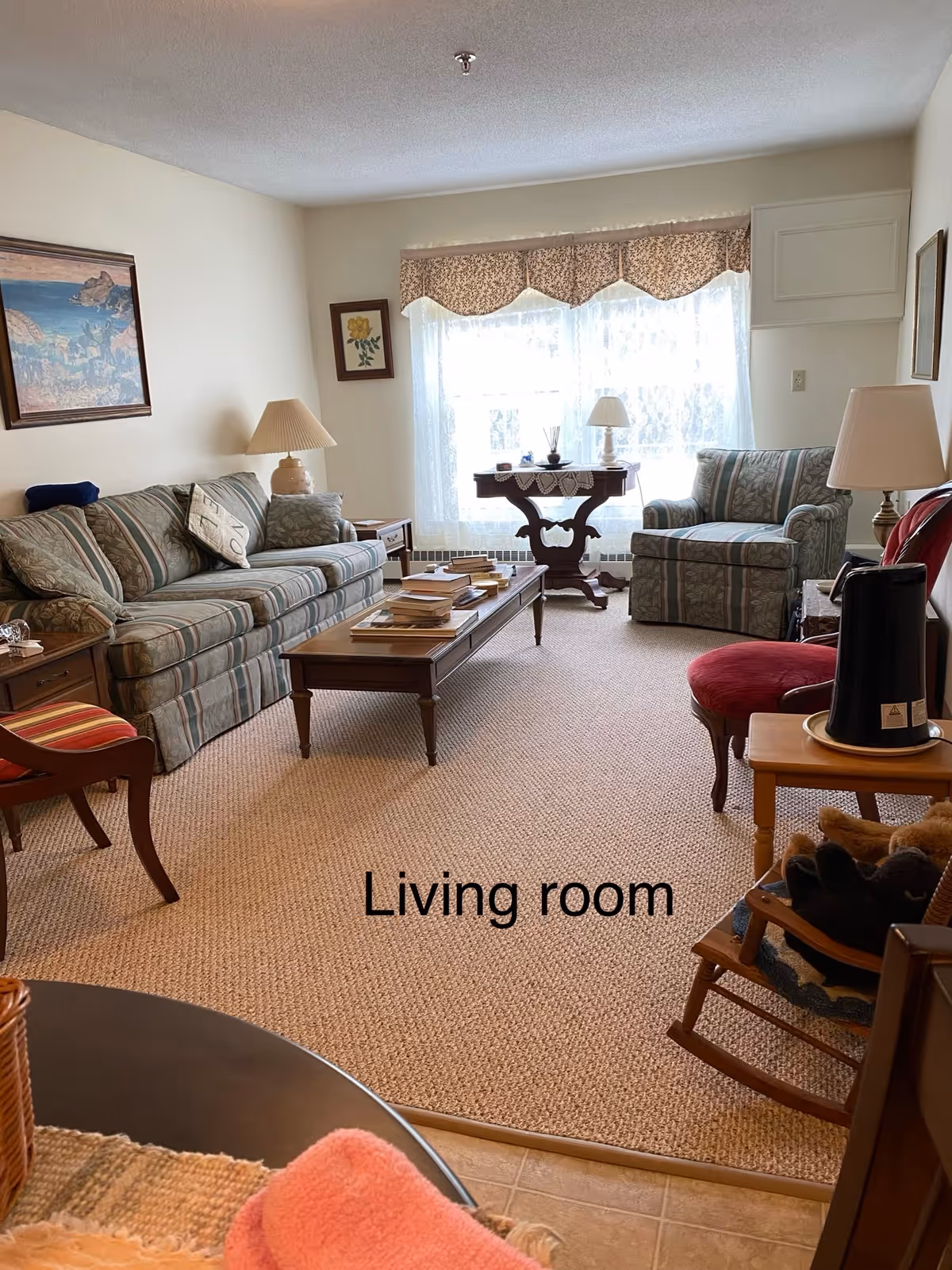 A cozy living room with a striped sofa, matching armchair, wooden coffee table with books, side tables with lamps, a rocking chair with stuffed animals, and a window with lace curtains and a floral valance.