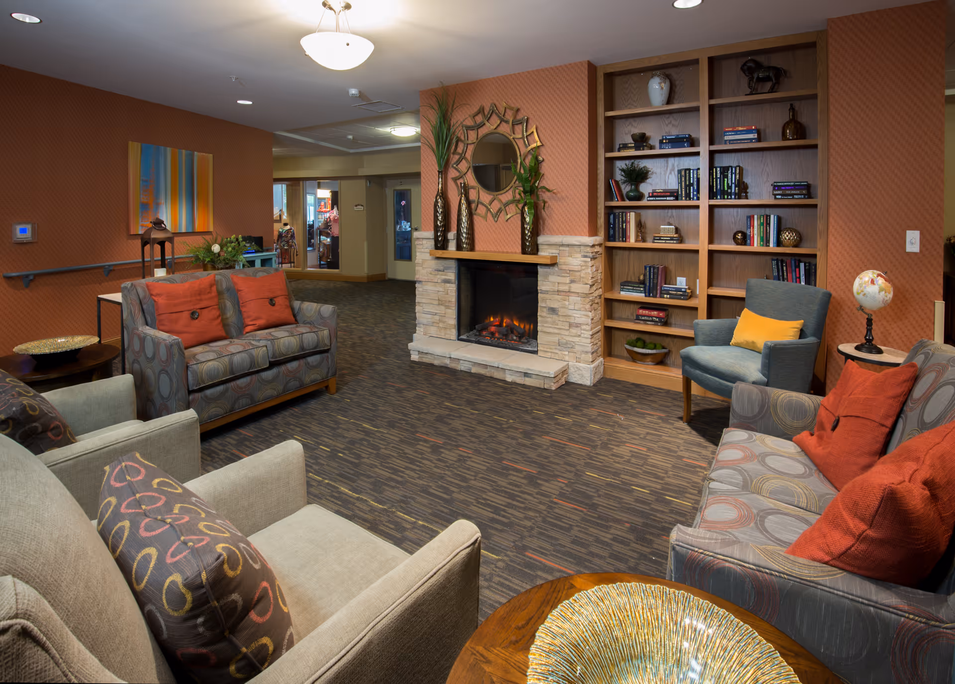 A cozy living room area in a senior living facility featuring a stone fireplace with a decorative mirror and vases above it. The room has multiple upholstered chairs and sofas with colorful cushions arranged around the fireplace. There is a wooden bookshelf filled with books and decorative items, and the walls are painted in warm tones with artwork and plants adding to the inviting atmosphere.