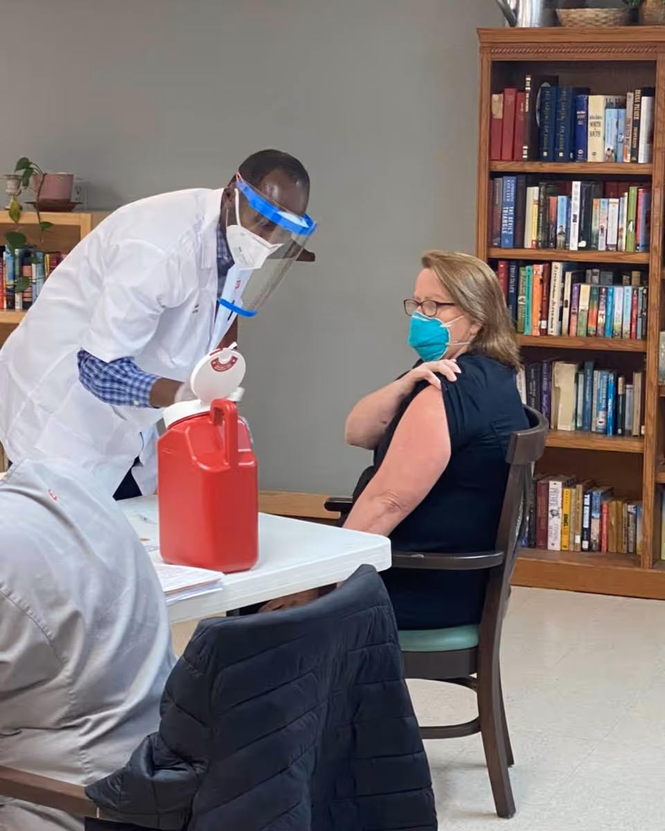 A healthcare worker wearing a white coat, face mask, and face shield is administering a vaccine to a seated woman wearing glasses and a face mask in a room with bookshelves filled with books.