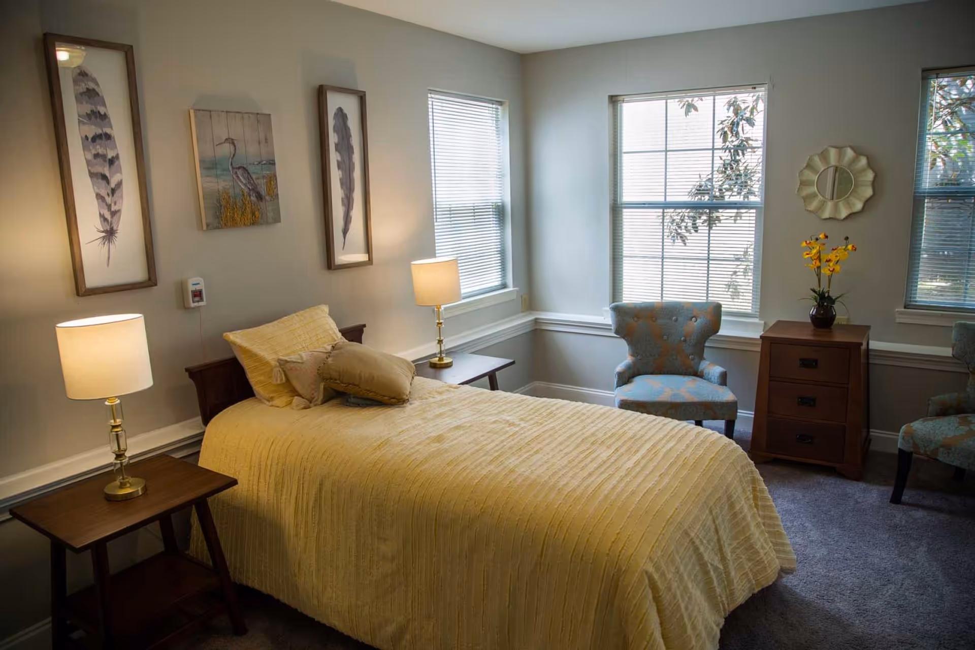 A cozy bedroom with a single bed covered in a yellow bedspread and multiple pillows. There are two wooden side tables with lamps on either side of the bed. The walls are decorated with framed artwork featuring feathers and a bird. Two windows with blinds allow natural light into the room. A wooden dresser with a vase of yellow flowers and a decorative mirror above it is positioned near two upholstered armchairs with a blue and orange pattern.