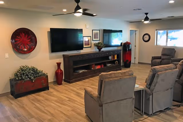 A cozy living room area with beige recliner chairs arranged facing a wall-mounted flat screen TV. The room features wood flooring, ceiling fans, decorative vases, framed pictures on the wall, a clock, and a large window letting in natural light.