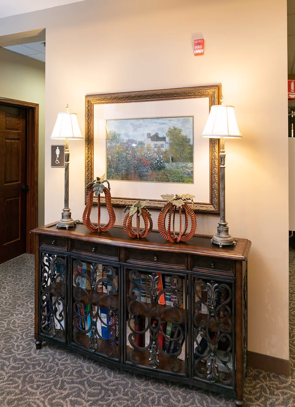 A decorative wooden cabinet with intricate metal doors, topped with two table lamps and three pumpkin-shaped ornaments. Above the cabinet hangs a framed painting of a garden scene with flowers and a house in the background. To the left, there is a wooden door with a sign indicating a women's restroom accessible for people with disabilities. The wall is painted beige and the floor is carpeted with a patterned design.