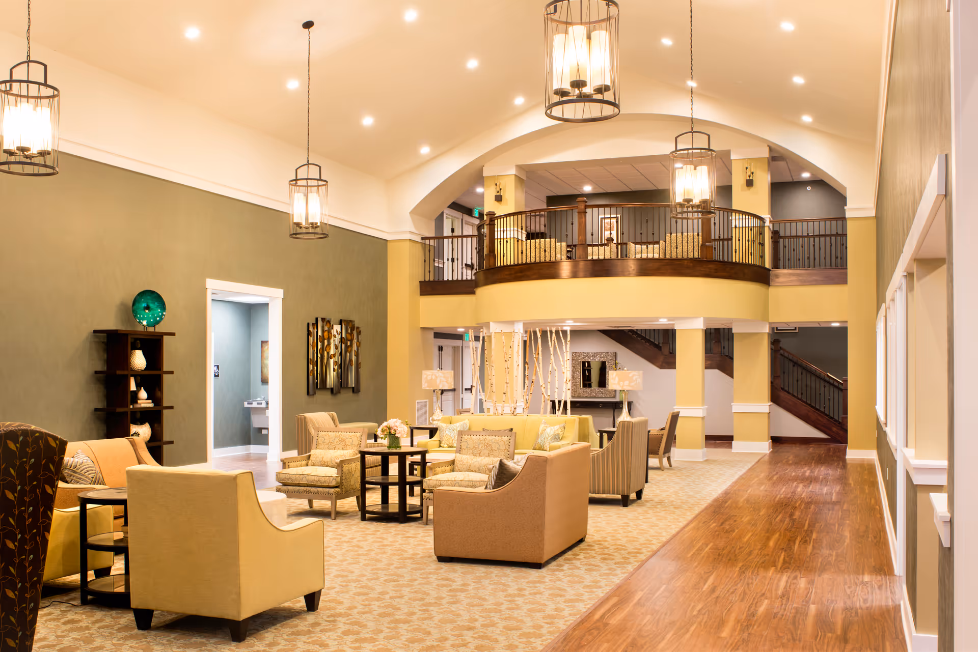 A spacious and well-lit living room area in a senior living facility with comfortable armchairs and sofas arranged around small tables. The room features high ceilings with hanging lantern-style lights, a mezzanine balcony with additional seating, and warm, neutral tones with green and beige walls. Hardwood flooring and carpeted areas complement the cozy and inviting atmosphere.