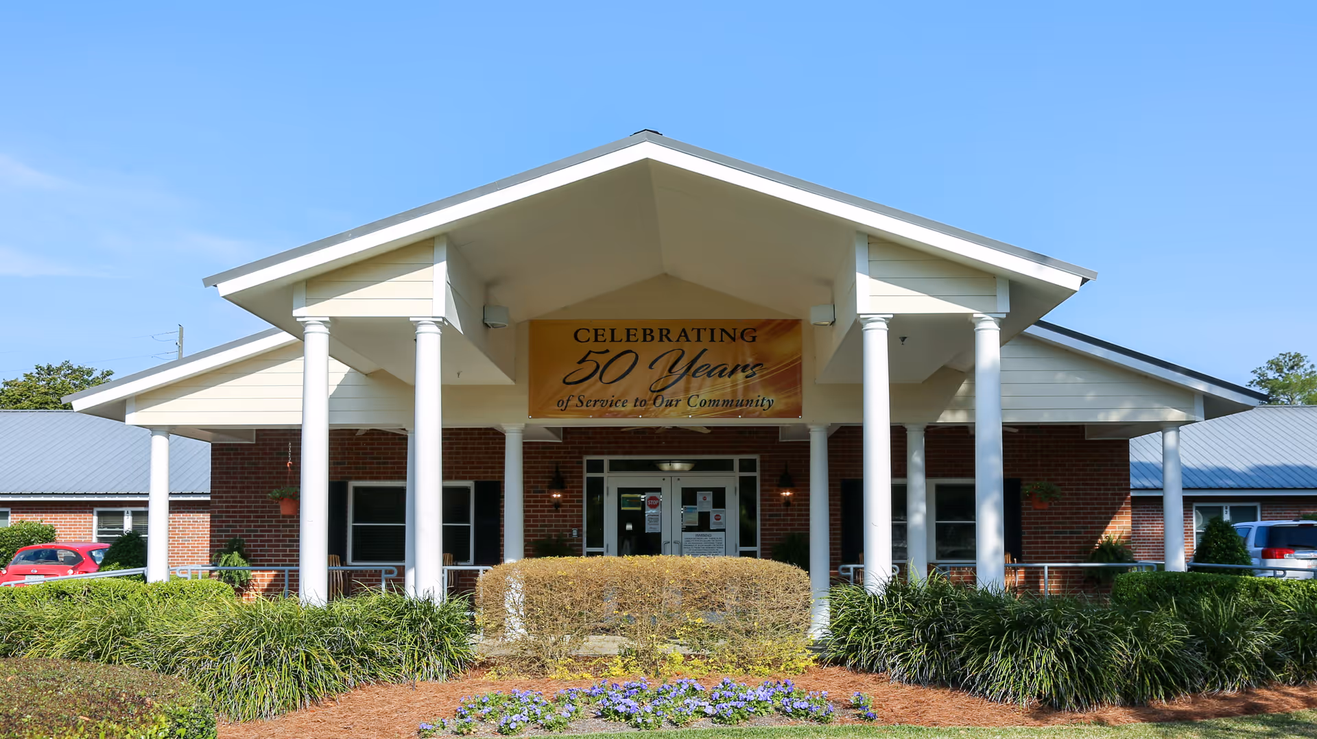 Front exterior view of Treutlen County Health & Rehabilitation building with a covered entrance supported by white columns. A banner above the entrance reads 'Celebrating 50 Years of Service to Our Community'. There are bushes and flowers in the landscaped area in front of the building, and cars are parked on either side.