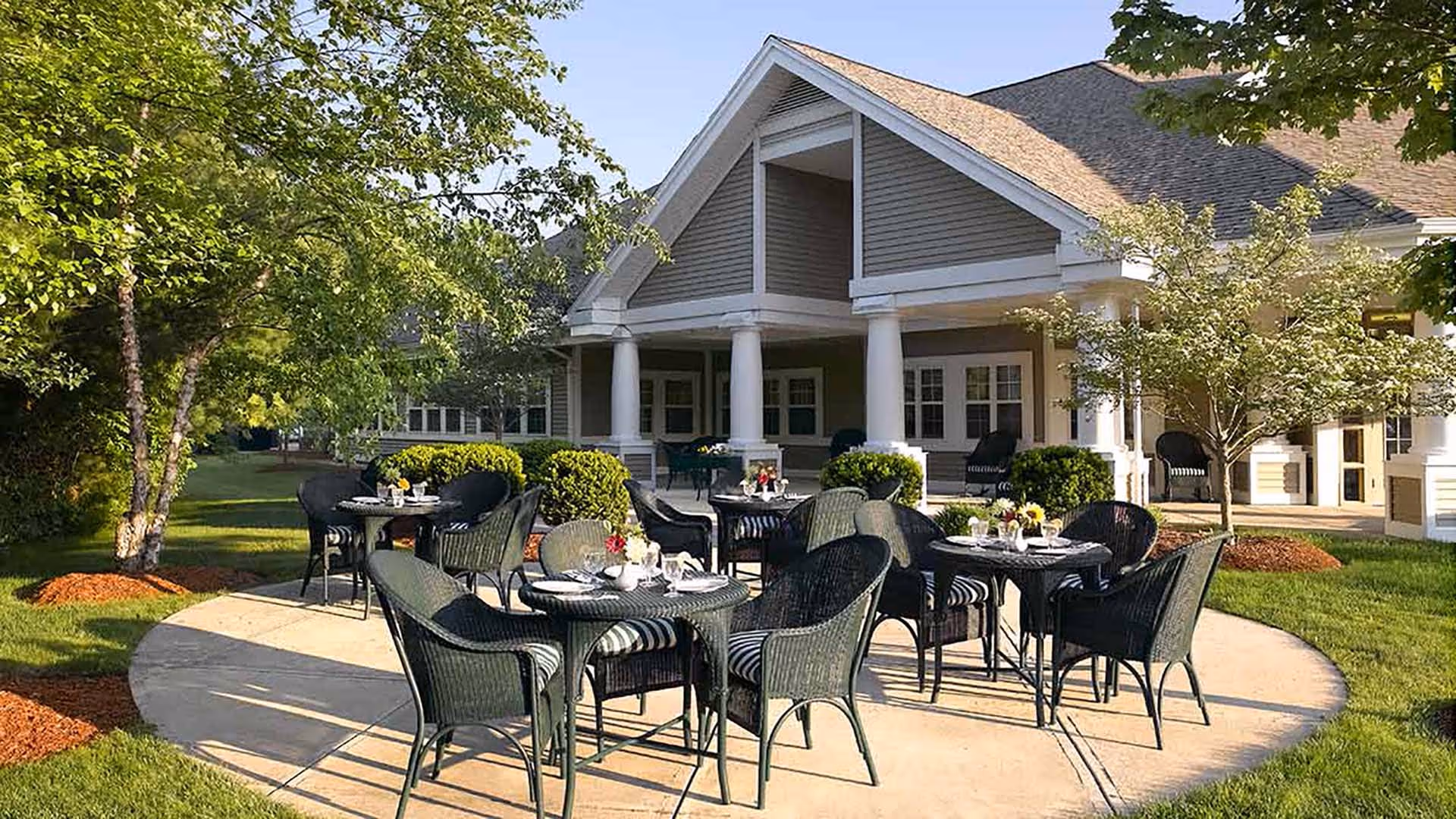 Outdoor patio area at Atria Kennebunk with several round tables and black wicker chairs arranged on a circular concrete surface. The patio is surrounded by green grass, trees, and shrubs, with a large building featuring white columns and multiple windows in the background.