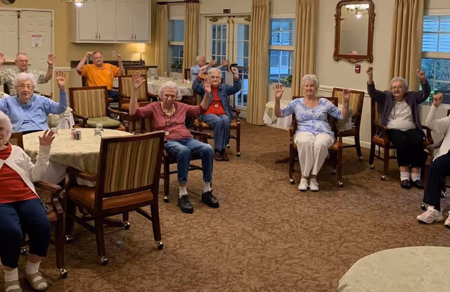 A group of elderly residents seated in a dining/activity room raising their hands during a group exercise.