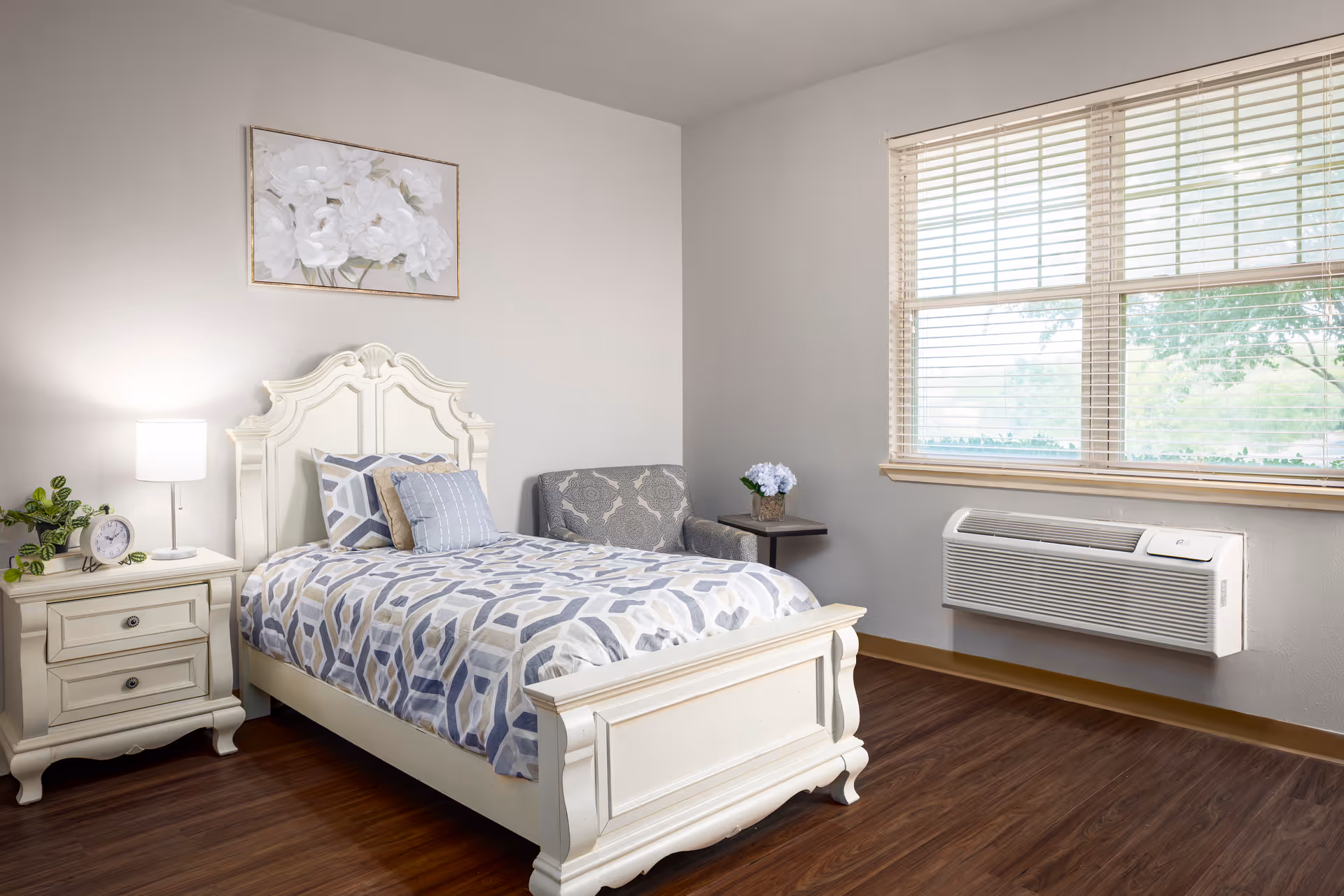 A bright and clean bedroom with a white wooden bed featuring a patterned blue and gray bedspread, a matching pillow, and a beige pillow. Next to the bed is a white nightstand with a lamp, a small plant, and a clock. There is a gray patterned armchair with a small side table holding a vase of flowers. A large window with blinds lets in natural light, and the floor is a dark wood laminate. A floral painting hangs on the wall above the bed.