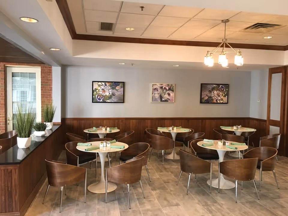 A dining room with four round tables, each set with green napkins and salt and pepper shakers. Each table is surrounded by four wooden chairs with metal legs. The room has a tiled floor, wood paneling on the lower half of the walls, and three floral paintings on the upper half. There are potted plants on a ledge by a window and a modern light fixture hanging from the ceiling.
