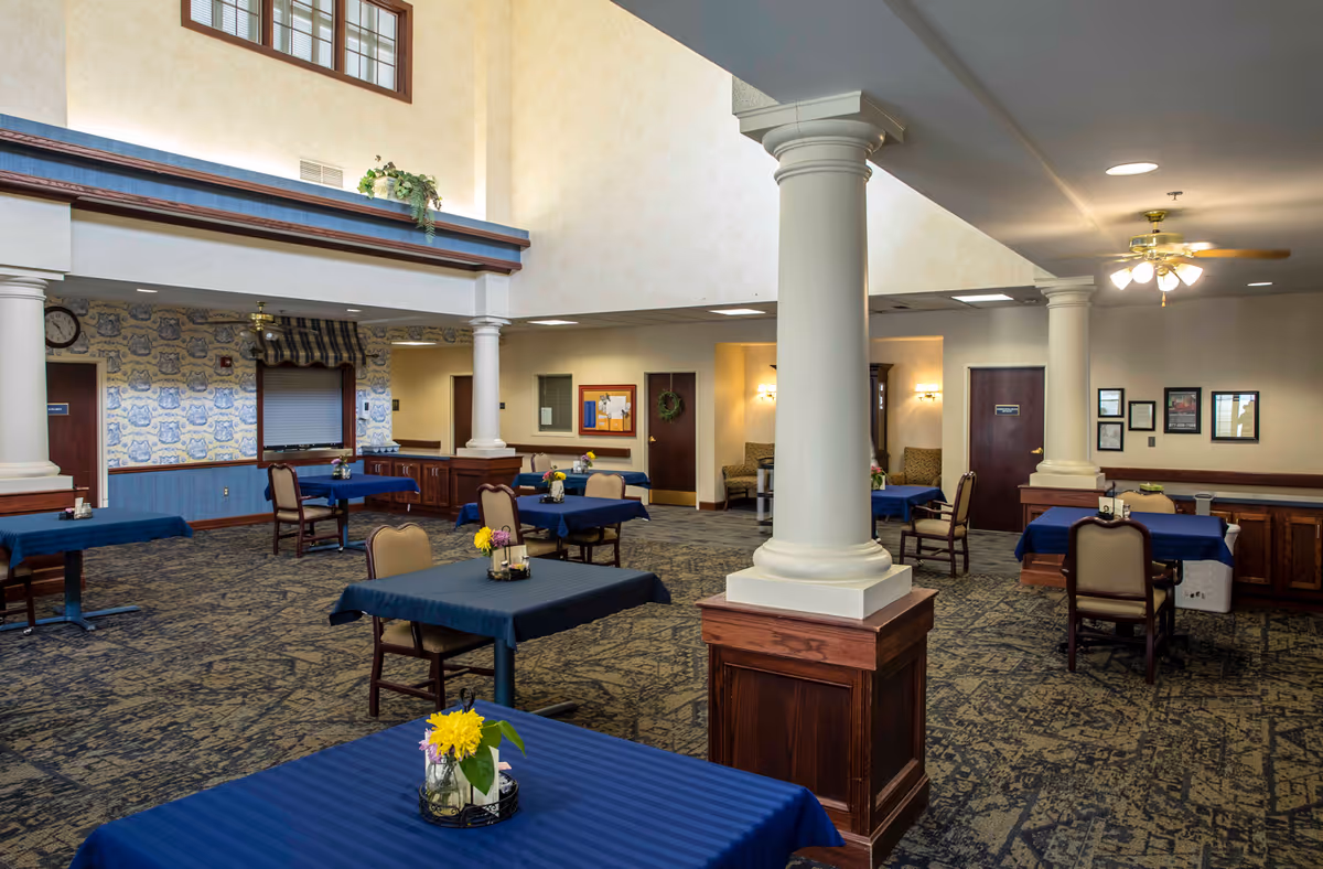 Spacious dining room with columns and multiple tables covered in blue tablecloths and small floral centerpieces.
