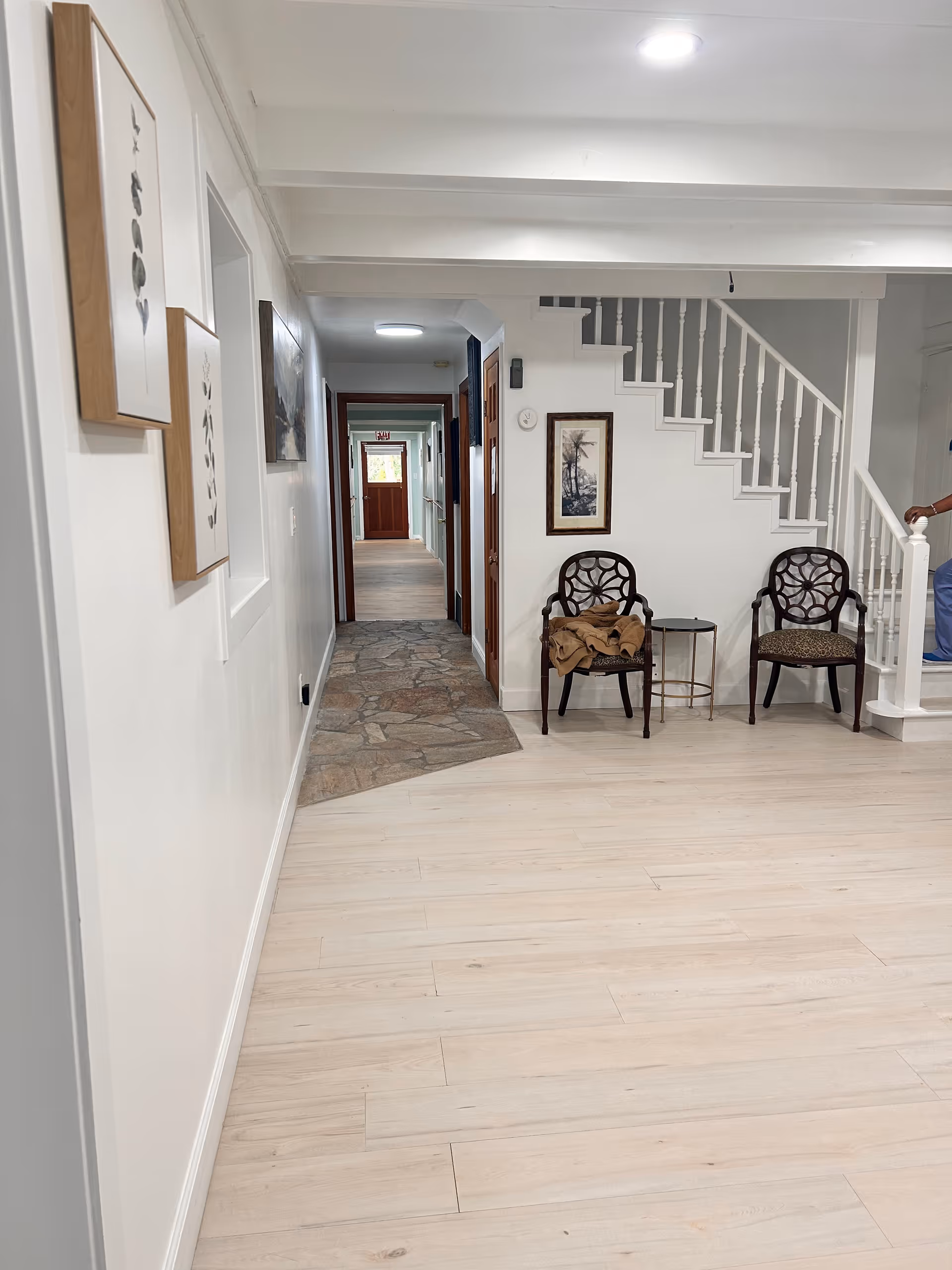 Interior view of a senior living facility hallway with light wood flooring and white walls. Two decorative chairs with a small round table are placed near a white staircase with wooden handrails. Artwork is hung on the walls, and a stone pathway leads down the hallway towards a wooden door with an exit sign above it.