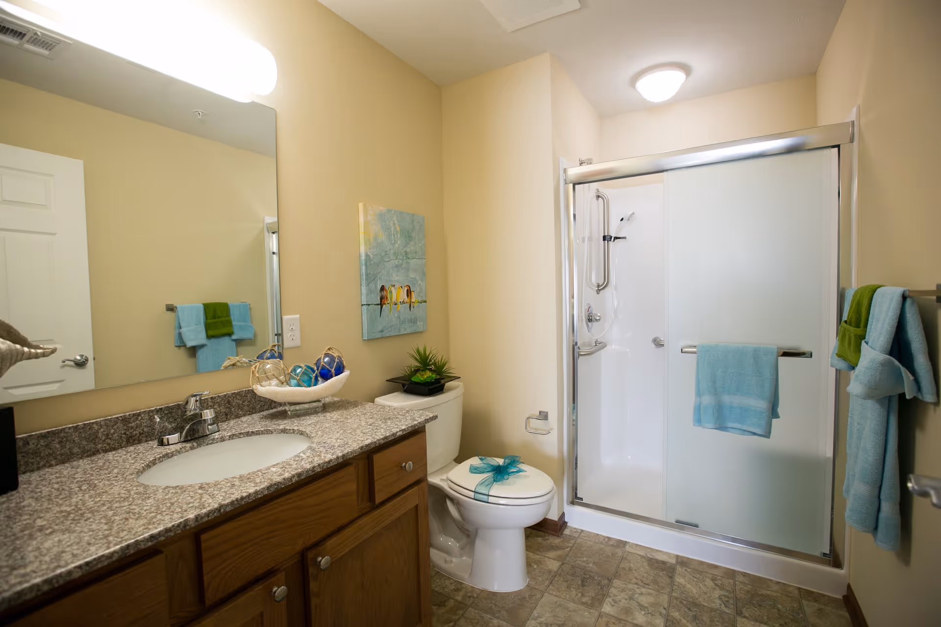 A clean and well-lit bathroom with a granite countertop vanity, a sink, a toilet with a decorative bow on the lid, and a shower with frosted glass sliding doors. The walls are painted beige, and there are blue and green towels hanging on racks. A small plant and decorative items are placed on the vanity and toilet tank. A painting of birds on a wire hangs on the wall above the toilet.
