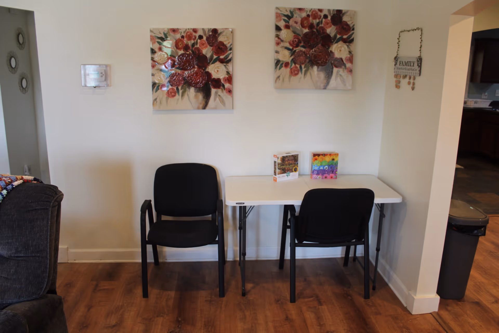 Two black chairs and a white folding table against a wall topped with floral paintings in a communal living area.