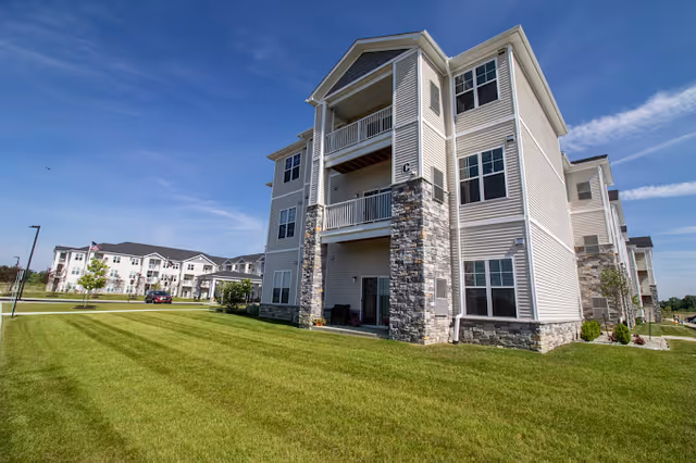 Three-story beige and stone-clad apartment building with balconies overlooking a wide green lawn under a blue sky.