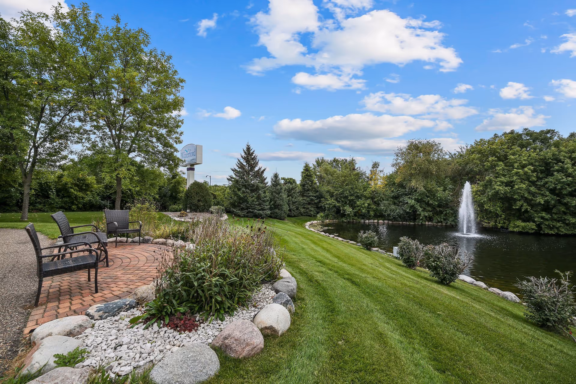 Landscaped outdoor seating area with chairs on a brick patio beside a pond with a fountain, surrounded by lawn and trees under a blue sky.