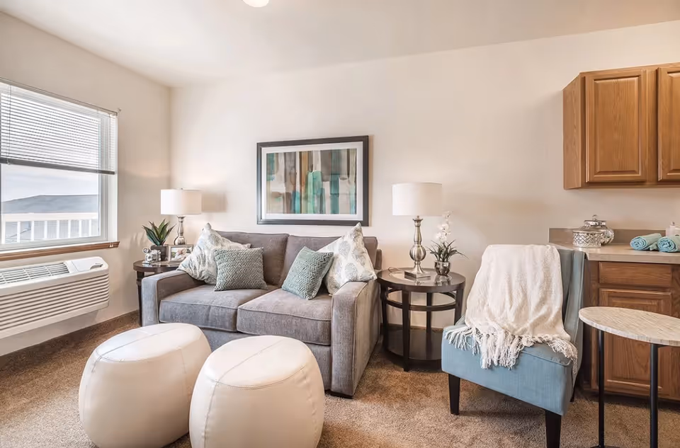 A cozy living room area with a gray sofa adorned with patterned and solid cushions, two white ottomans in front, a blue armchair draped with a white throw blanket, two round side tables with lamps and decorative plants, a framed abstract painting on the wall, a window with blinds, and wooden kitchen cabinets on the right.
