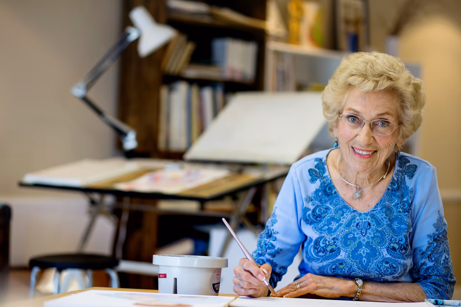 An elderly woman with short curly blonde hair and glasses is smiling while painting at a table. She is wearing a blue patterned top and is holding a paintbrush. In the background, there is a bookshelf with books and an adjustable desk lamp.