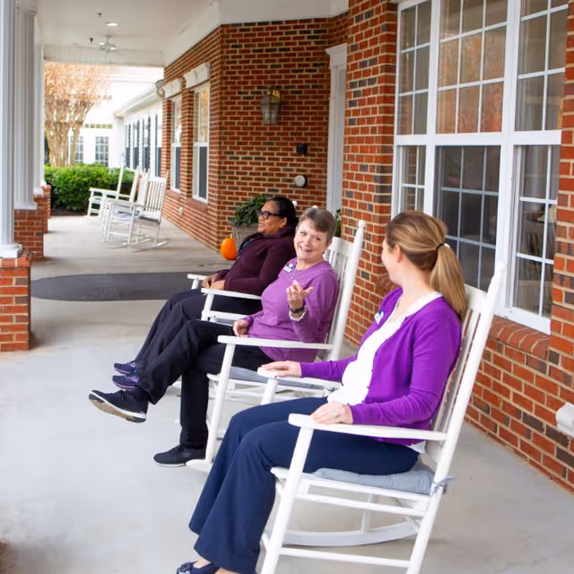 Three women sitting and chatting on white rocking chairs on a covered porch with brick walls and large windows at a senior living facility.