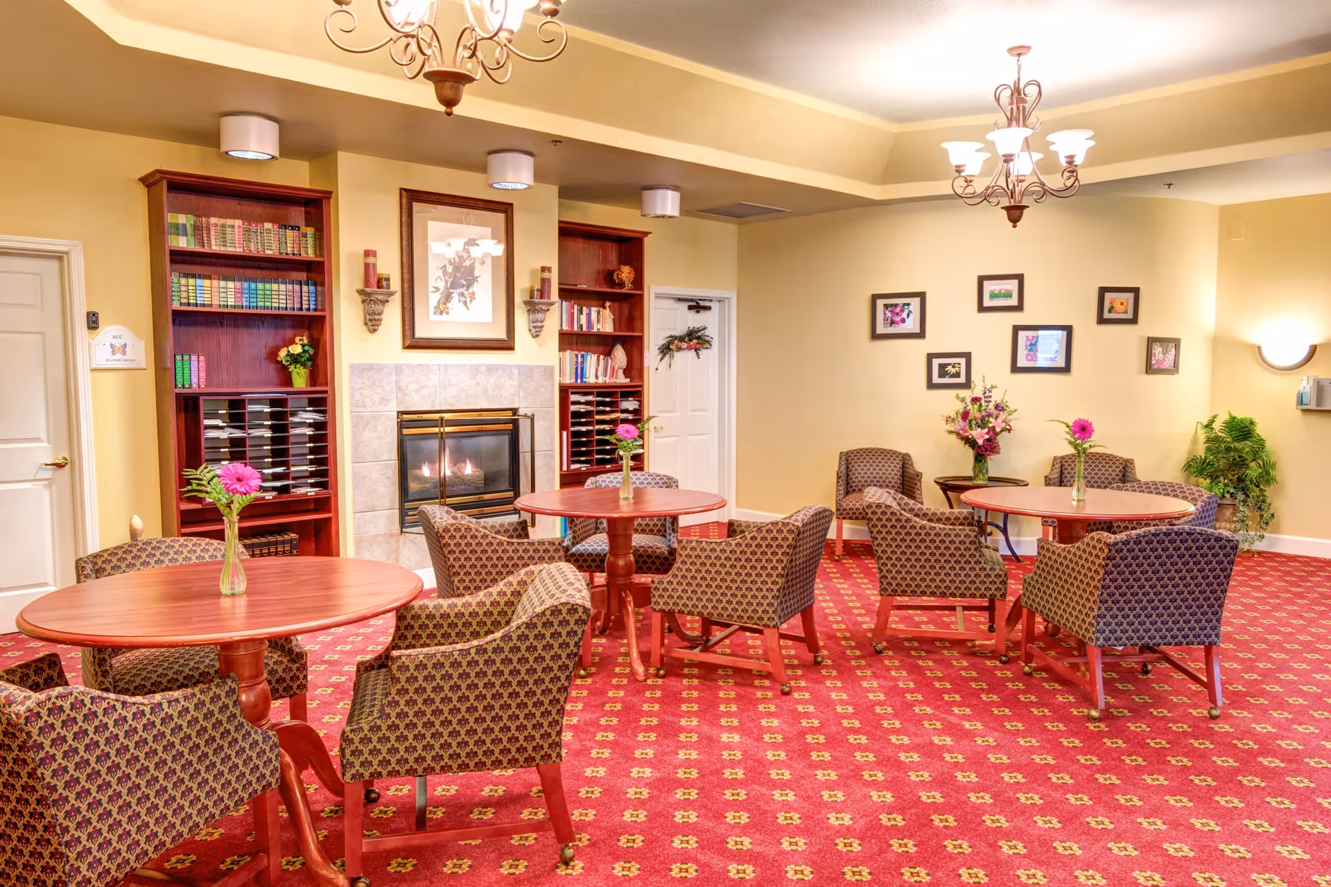 A cozy common area with patterned armchairs and round wooden tables, each table decorated with a small vase holding a single flower. The room features a red carpet with a floral pattern, a fireplace with a tiled surround, built-in wooden bookshelves filled with books and decorative items, and framed artwork on the walls. Two chandeliers hang from the ceiling, providing warm lighting.