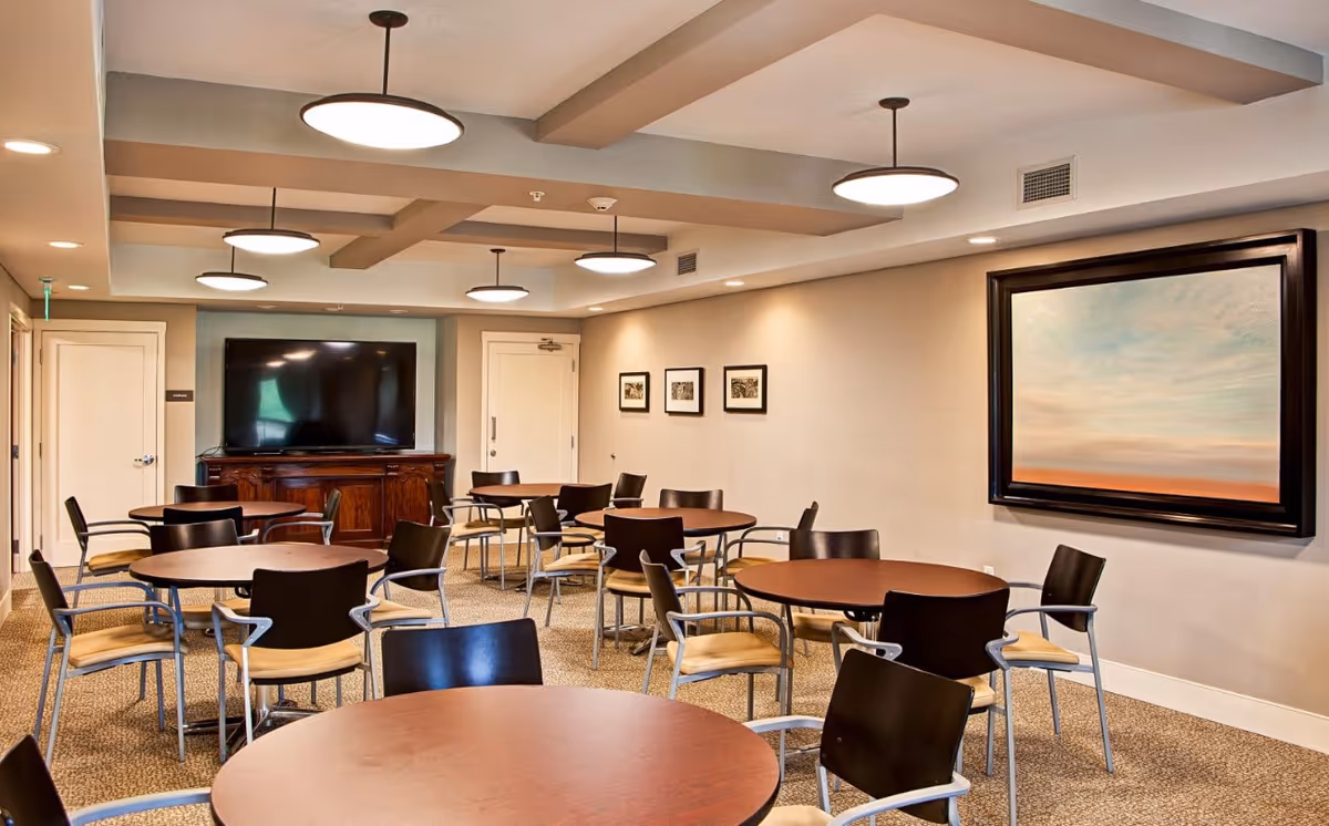 A well-lit common room with multiple round tables and chairs arranged for group seating. The room features a large flat-screen TV mounted on a wooden cabinet against the far wall, neutral-colored walls with framed artwork, and modern ceiling lights.