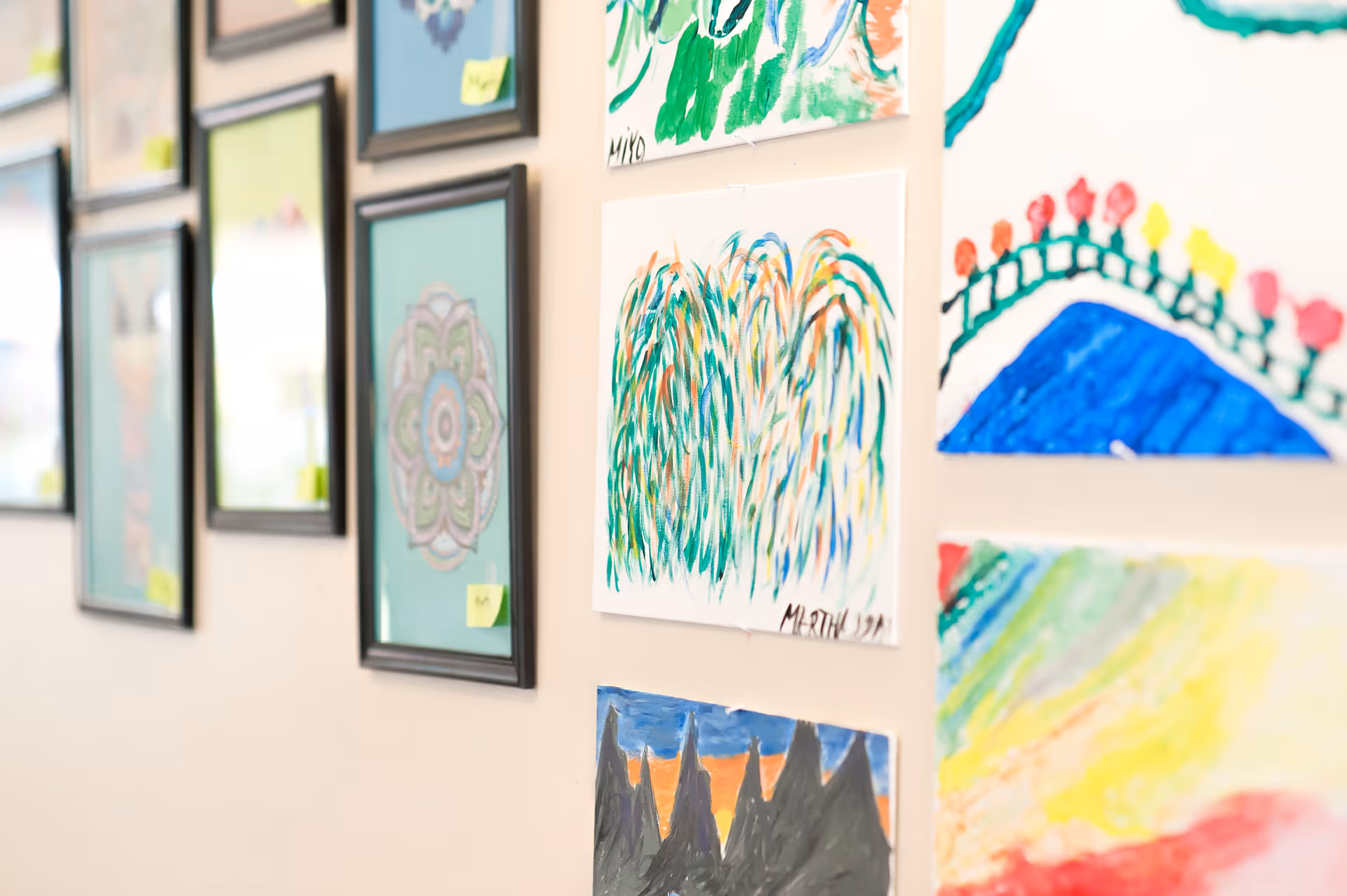 A wall decorated with various framed artworks and colorful paintings, including abstract designs and nature scenes, displayed in a senior living facility.