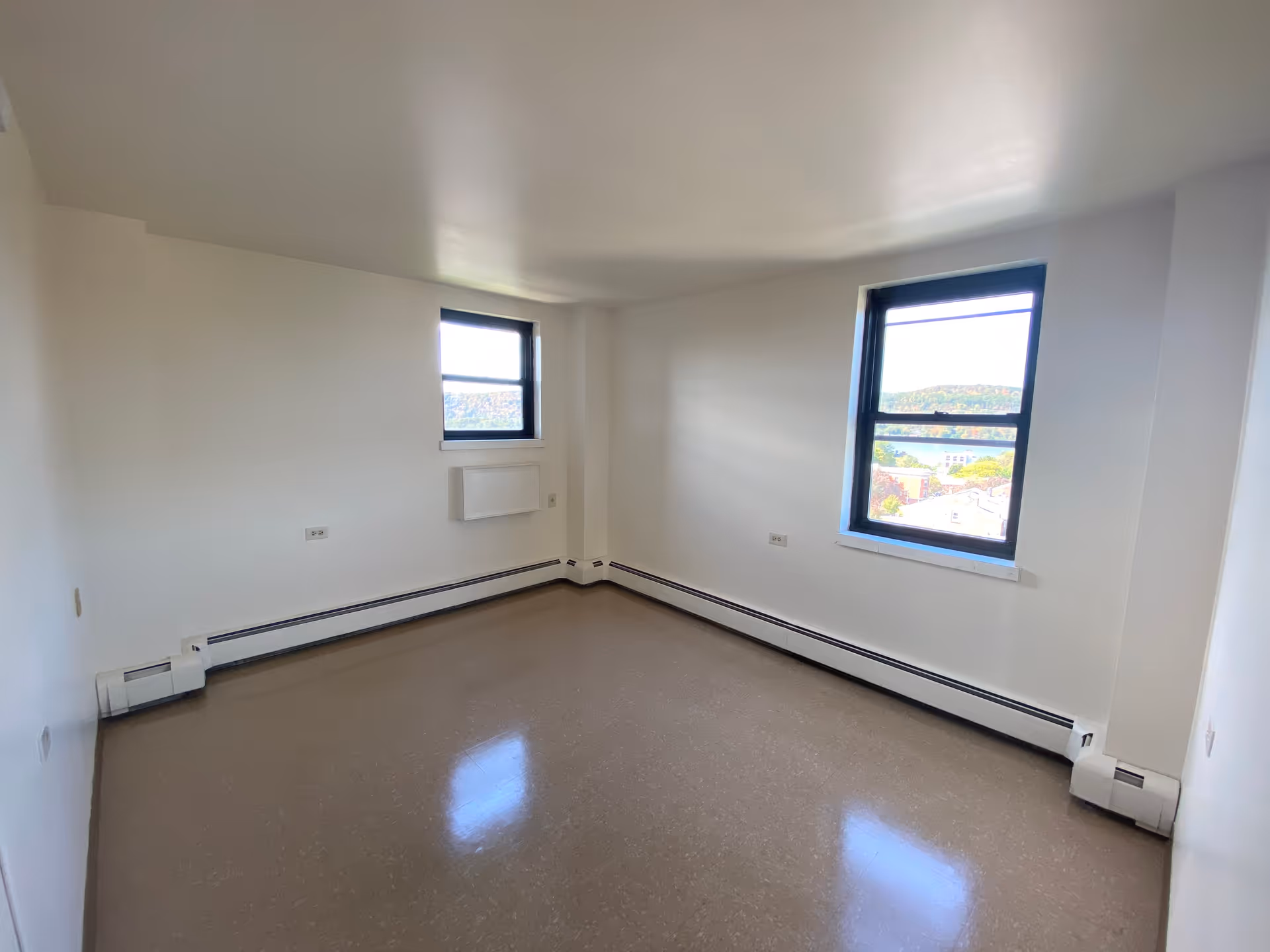 Empty room with two windows showing an outdoor view, white walls, and brown flooring with baseboard heating along the walls.