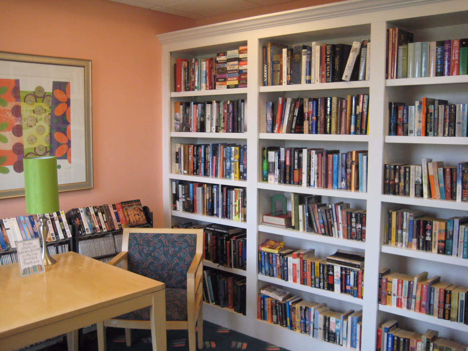 A cozy reading area with a wooden table, a chair with patterned upholstery, a green lamp, and a large white bookshelf filled with books against a peach-colored wall. There is also a colorful framed artwork on the wall and a small rack with more books or media.