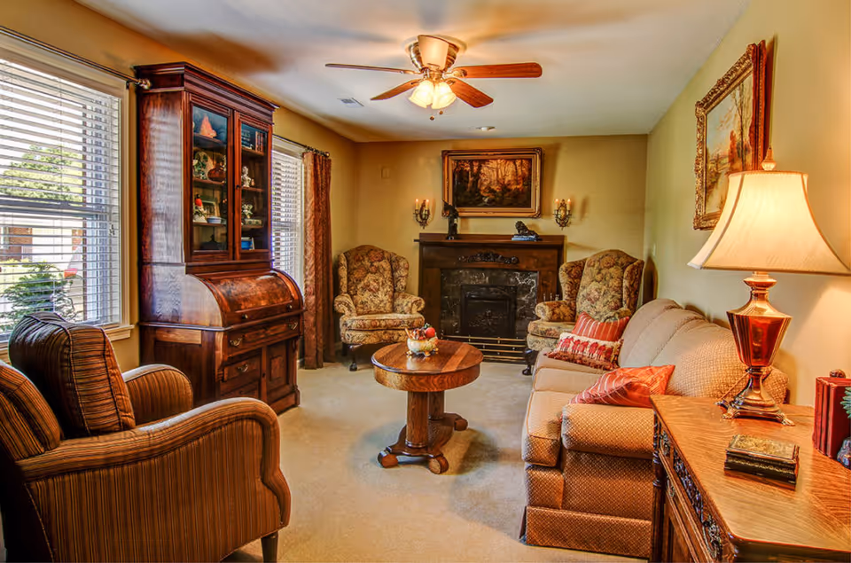 A cozy living room with a beige sofa adorned with red and patterned pillows, two floral upholstered armchairs, a wooden round coffee table with a decorative bowl, and a wooden cabinet with glass doors. The room features a fireplace with a dark marble surround, a painting above it, wall sconces with candles, a ceiling fan with lights, and a table lamp on a wooden side table. Large windows with blinds and curtains let in natural light.