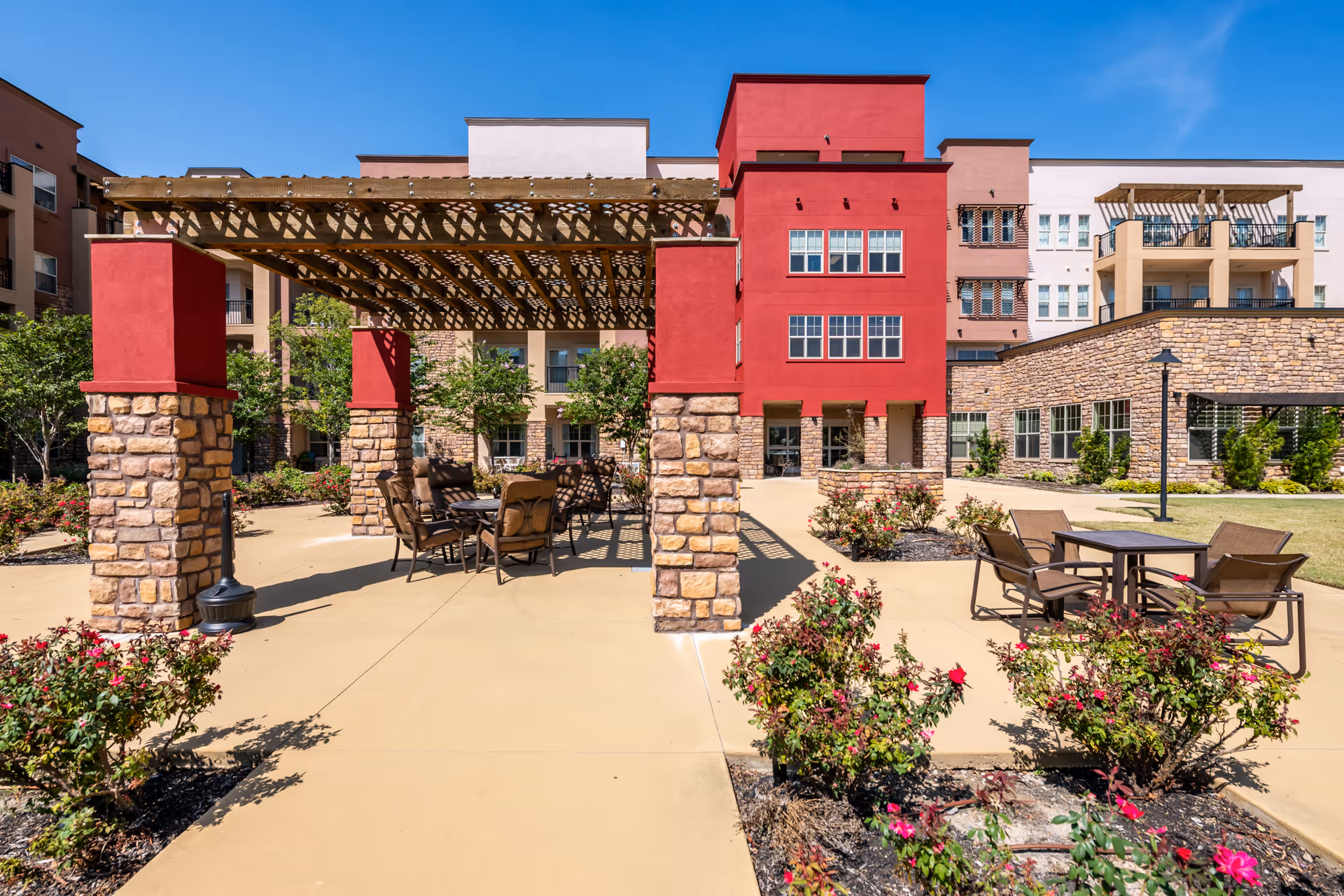 Outdoor patio area at Alexis Estates Gracious Retirement Living featuring a pergola with stone and red pillars, several tables and chairs for seating, surrounded by landscaped flower beds with blooming pink flowers, and a multi-story building in the background under a clear blue sky.