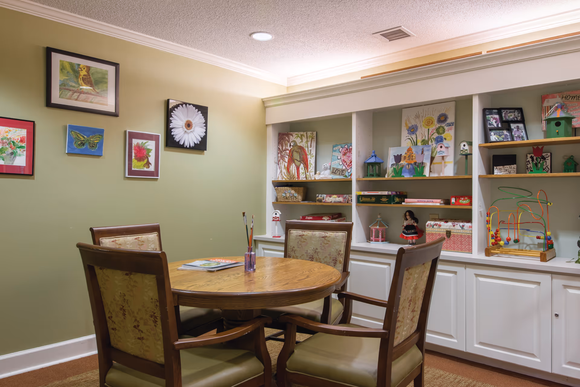 Round wooden table with four upholstered chairs in a cozy activity room with built-in shelves displaying crafts and artwork.