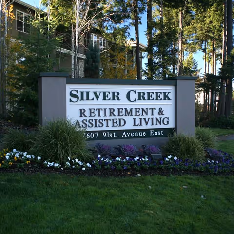 Outdoor view of a large sign for Silver Creek Retirement & Assisted Living, surrounded by well-maintained landscaping with flowers and greenery, with trees and part of a building visible in the background.