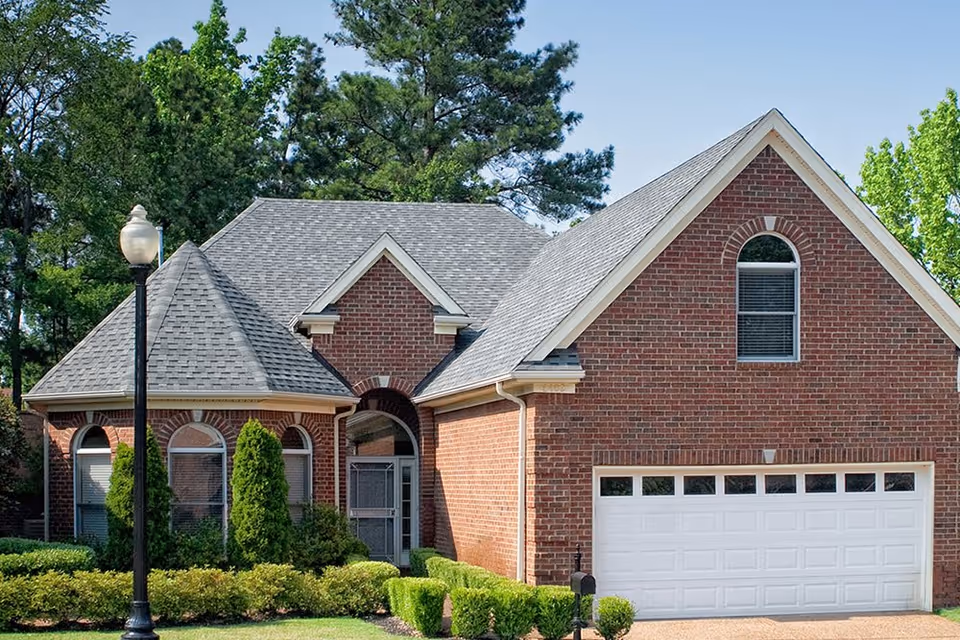 A brick residential building with a gray shingled roof, featuring a two-car white garage door, arched windows, and a small front garden with trimmed bushes and a street lamp.