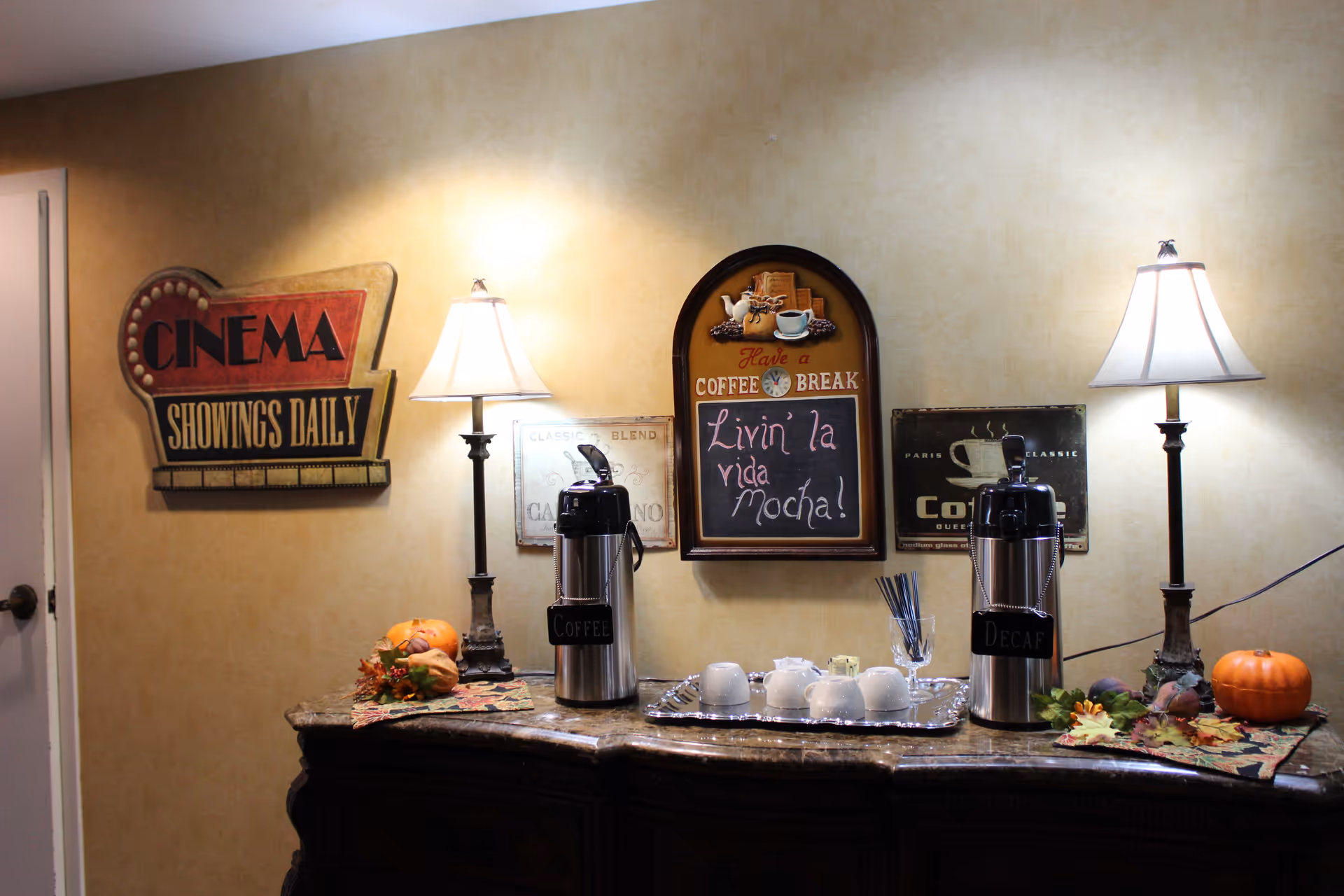 A decorated indoor coffee station with two beverage dispensers, lamps, cups on a tray, pumpkins, and wall signs including a chalkboard reading "Livin' la vida mocha".