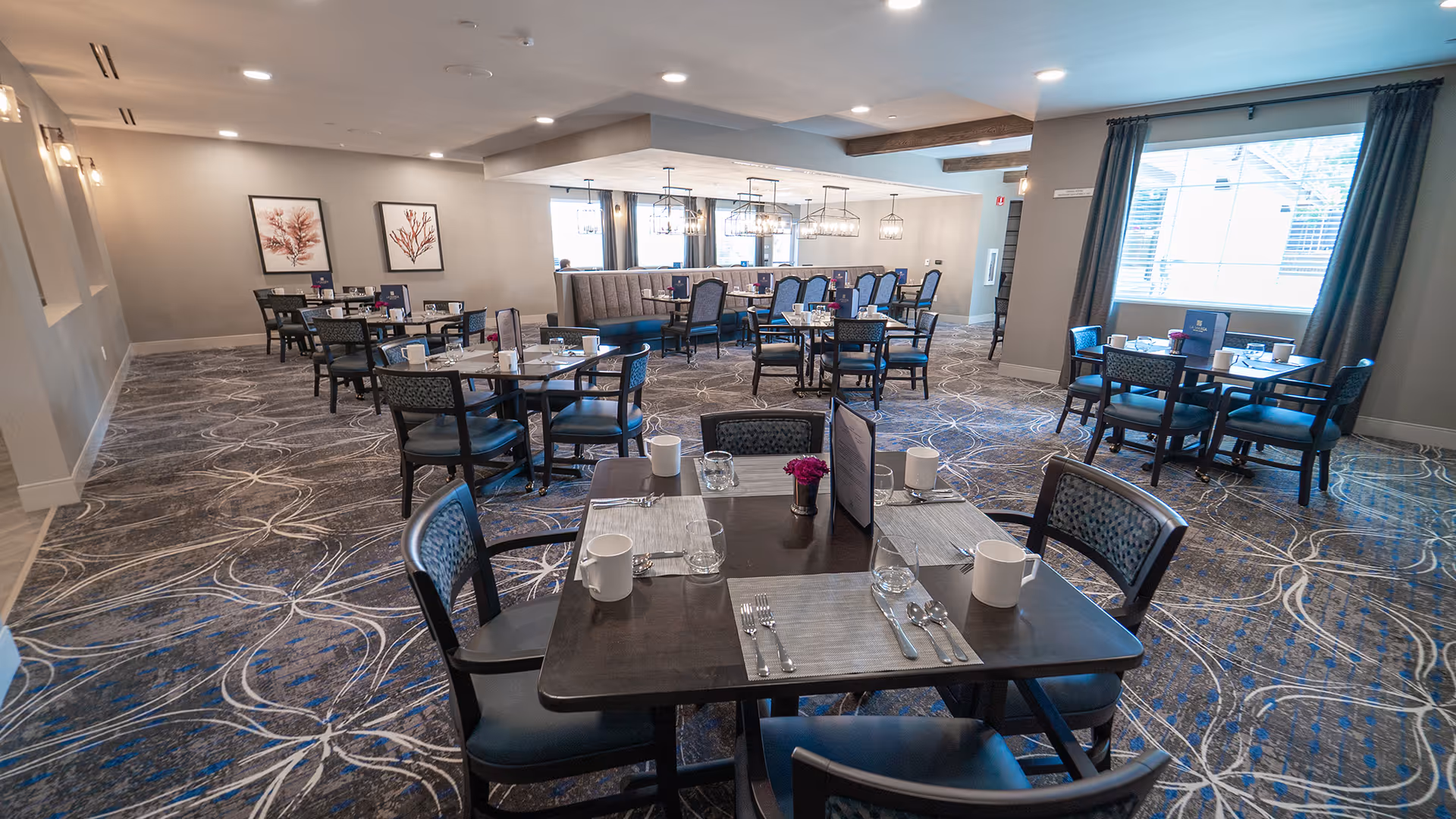 Spacious senior living dining room with multiple set tables and chairs, patterned carpet, and large windows.