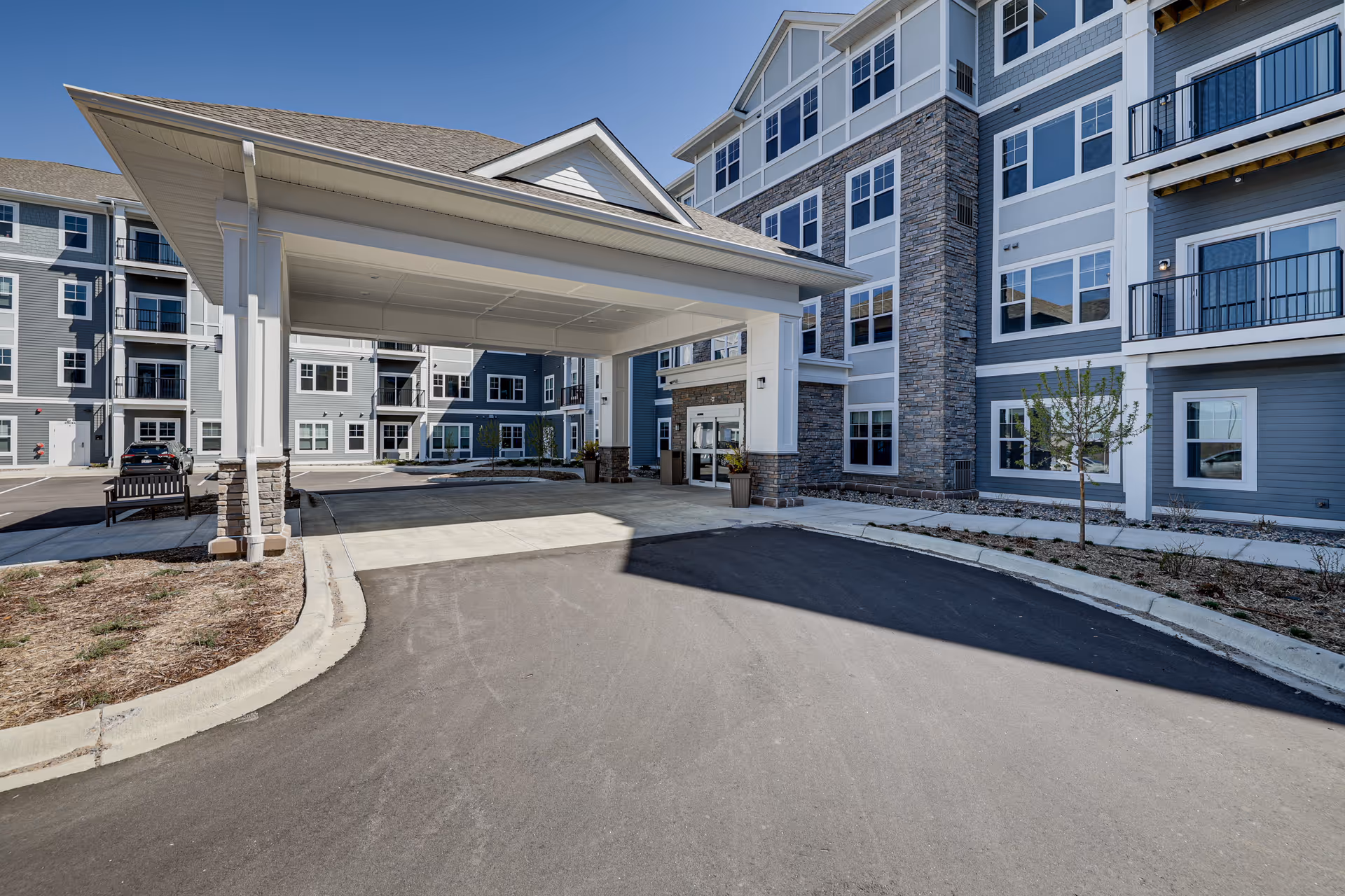 Entrance of a multi-story senior living facility with a covered drop-off area, stone and siding exterior, and multiple windows. There is a paved driveway and some landscaping with small trees and shrubs.