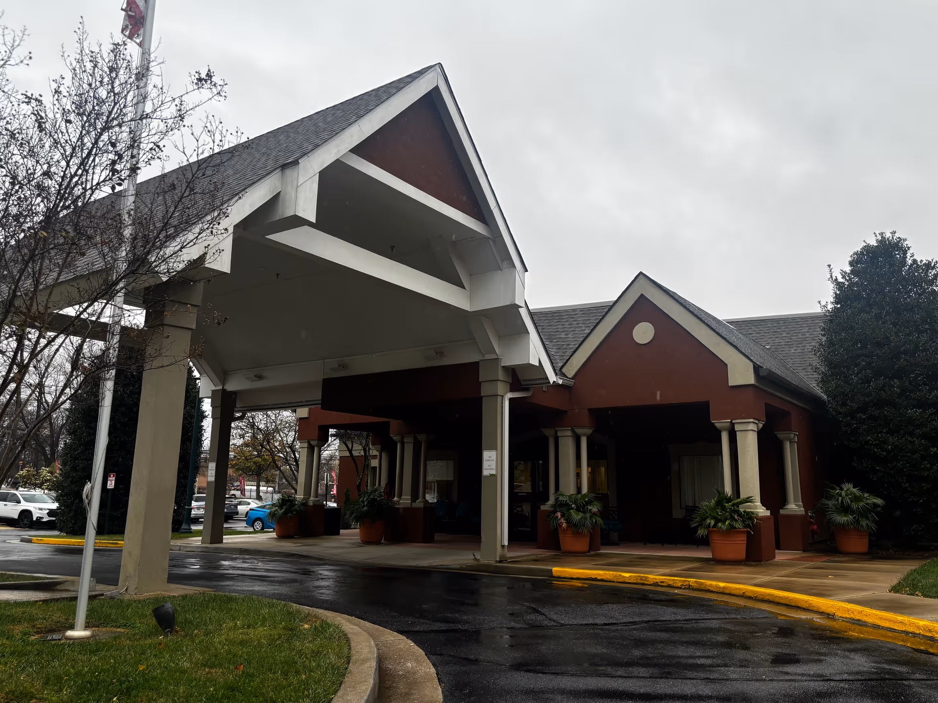 Exterior view of Aspenwood Senior Living Community building entrance with a covered driveway, potted plants, and a flagpole on a cloudy day.