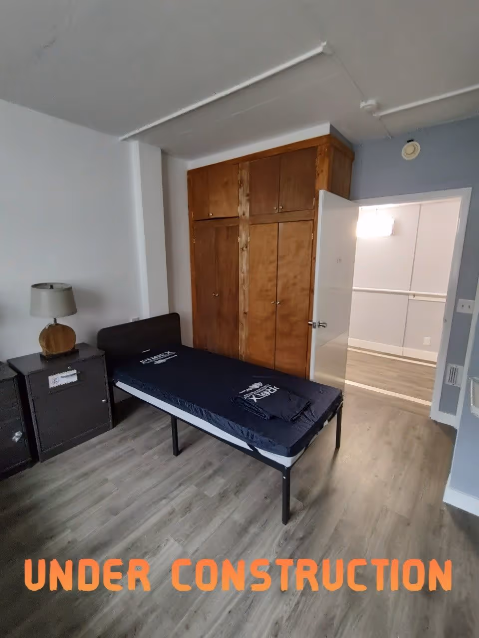 A small bedroom with a single bed frame and mattress, a wooden nightstand with a lamp, a large wooden built-in closet, and an open door leading to a hallway. The floor is covered with wood-like laminate, and the room appears to be under construction or renovation as indicated by the text 'UNDER CONSTRUCTION' at the bottom of the image.