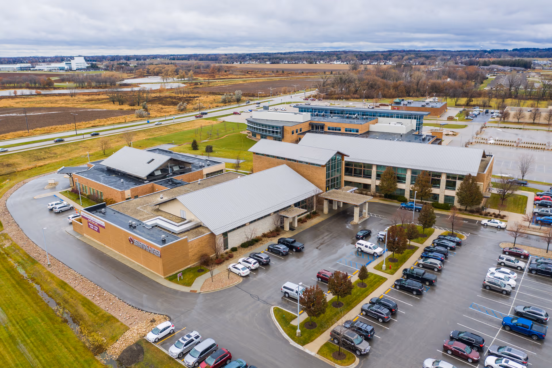 Aerial view of a large senior living facility named Recovery Inn with multiple connected buildings, a covered entrance, and a parking lot filled with cars. Surrounding the facility are open fields, roads, and some trees with autumn foliage.
