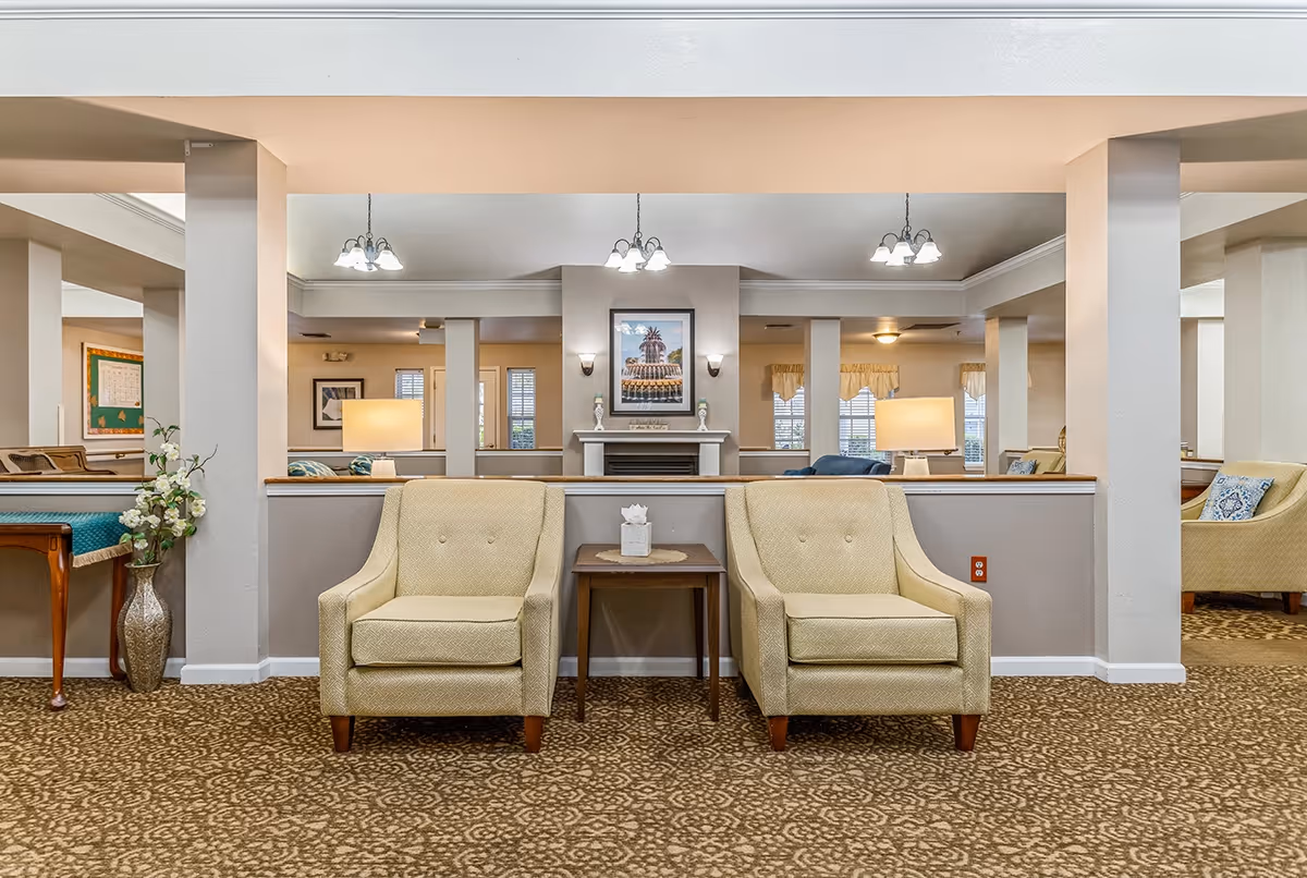 A cozy senior living facility common area with two beige armchairs separated by a small wooden table holding a tissue box. The room features a patterned carpet, a fireplace with a framed picture above it, and multiple light fixtures hanging from the ceiling. There are additional seating areas and windows with curtains in the background.