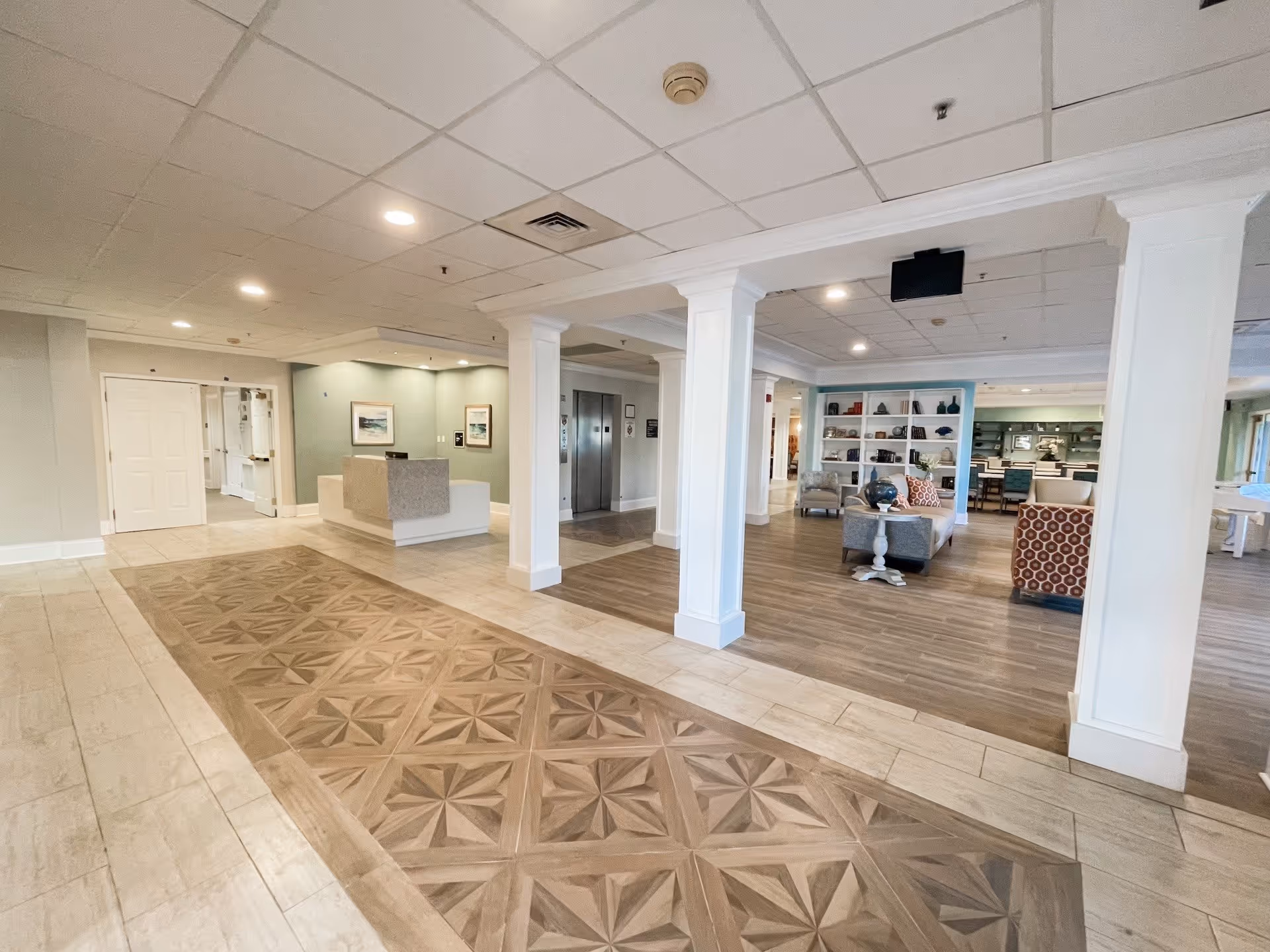 Interior view of a senior living facility lobby area with a reception desk on the left, two elevators in the center, and a seating area with chairs and a bookshelf on the right. The floor has a decorative tile pattern in the middle, and the ceiling has recessed lighting.