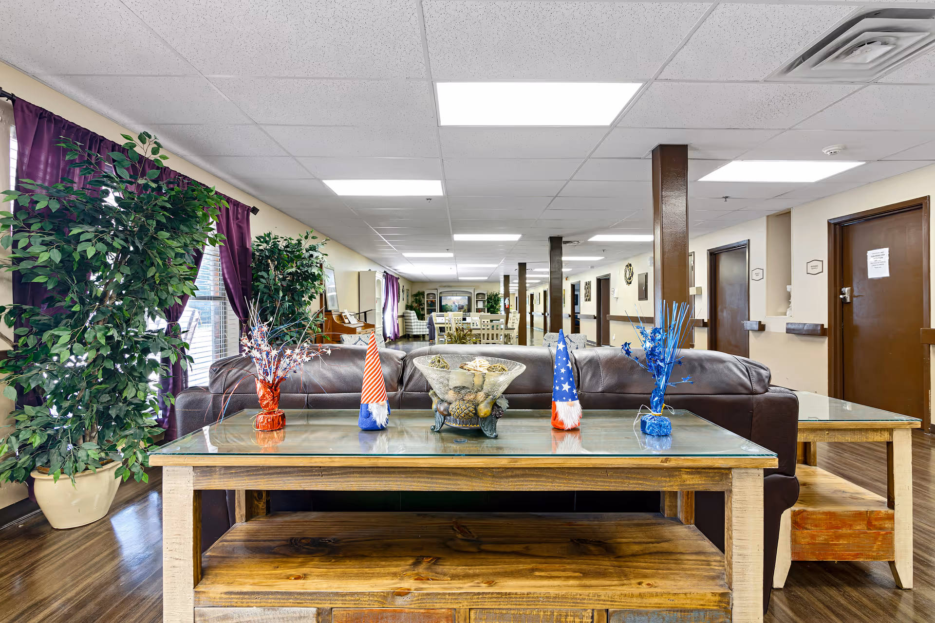 Interior view of a nursing and rehab facility lounge area with a brown leather couch, a wooden coffee table with glass top decorated with patriotic-themed ornaments and a bowl of decorative items. The room has wooden floors, potted plants, purple curtains on windows, and a long hallway with multiple doors and seating areas visible in the background.