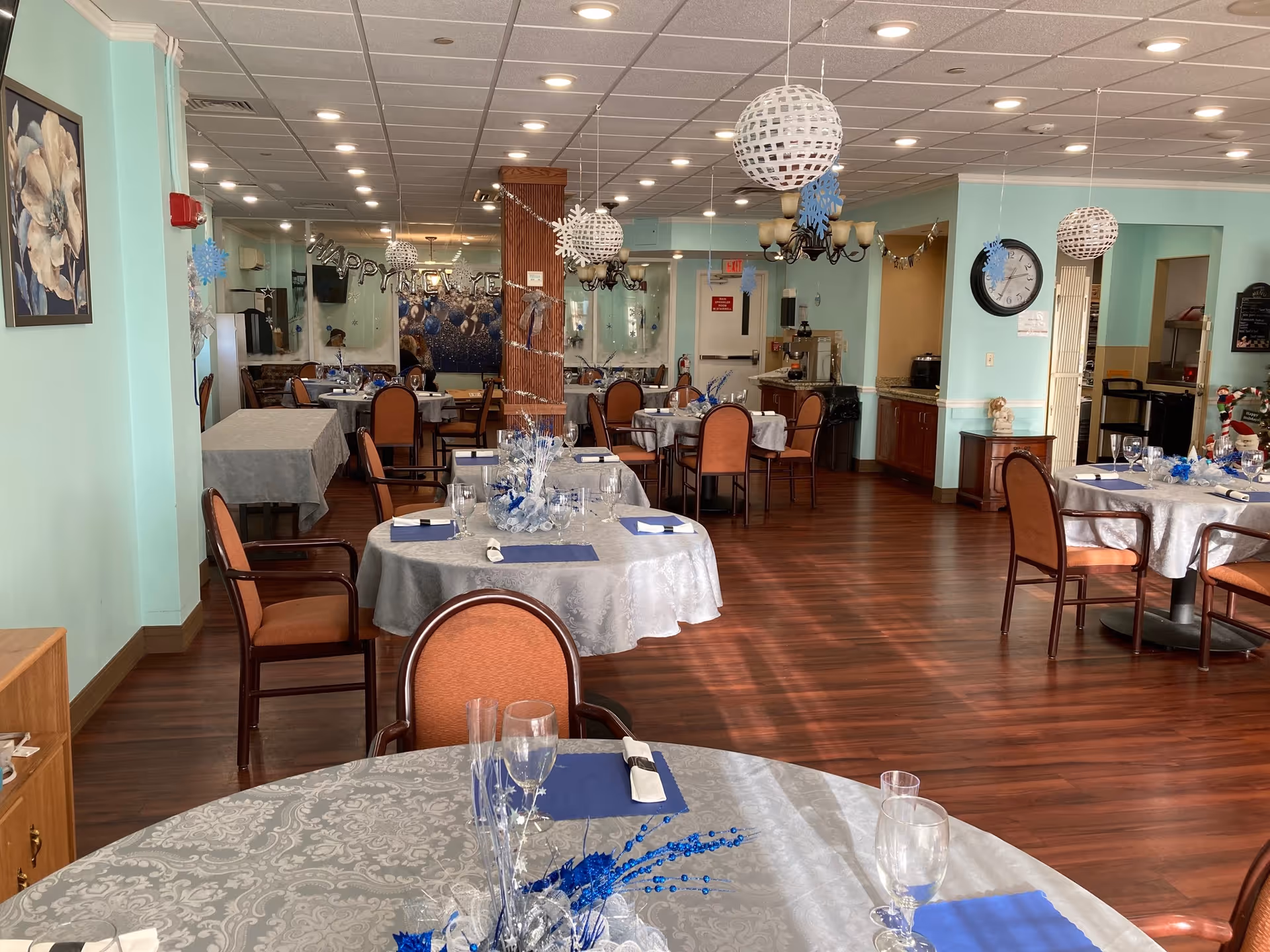 A dining room in St. Germain Manor decorated for a celebration with round tables covered in white patterned tablecloths, blue napkins, and glassware. The room has wooden floors, light blue walls, and hanging white spherical decorations with snowflakes. There are several chairs around each table and a clock on the wall near a doorway.