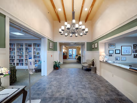 Interior view of a senior living facility hallway with a tiled floor, a modern chandelier hanging from a ceiling with wooden beams, and a reception desk on the right. To the left, there is an open doorway leading to a room with bookshelves filled with books. The hallway extends forward with seating areas and plants visible.